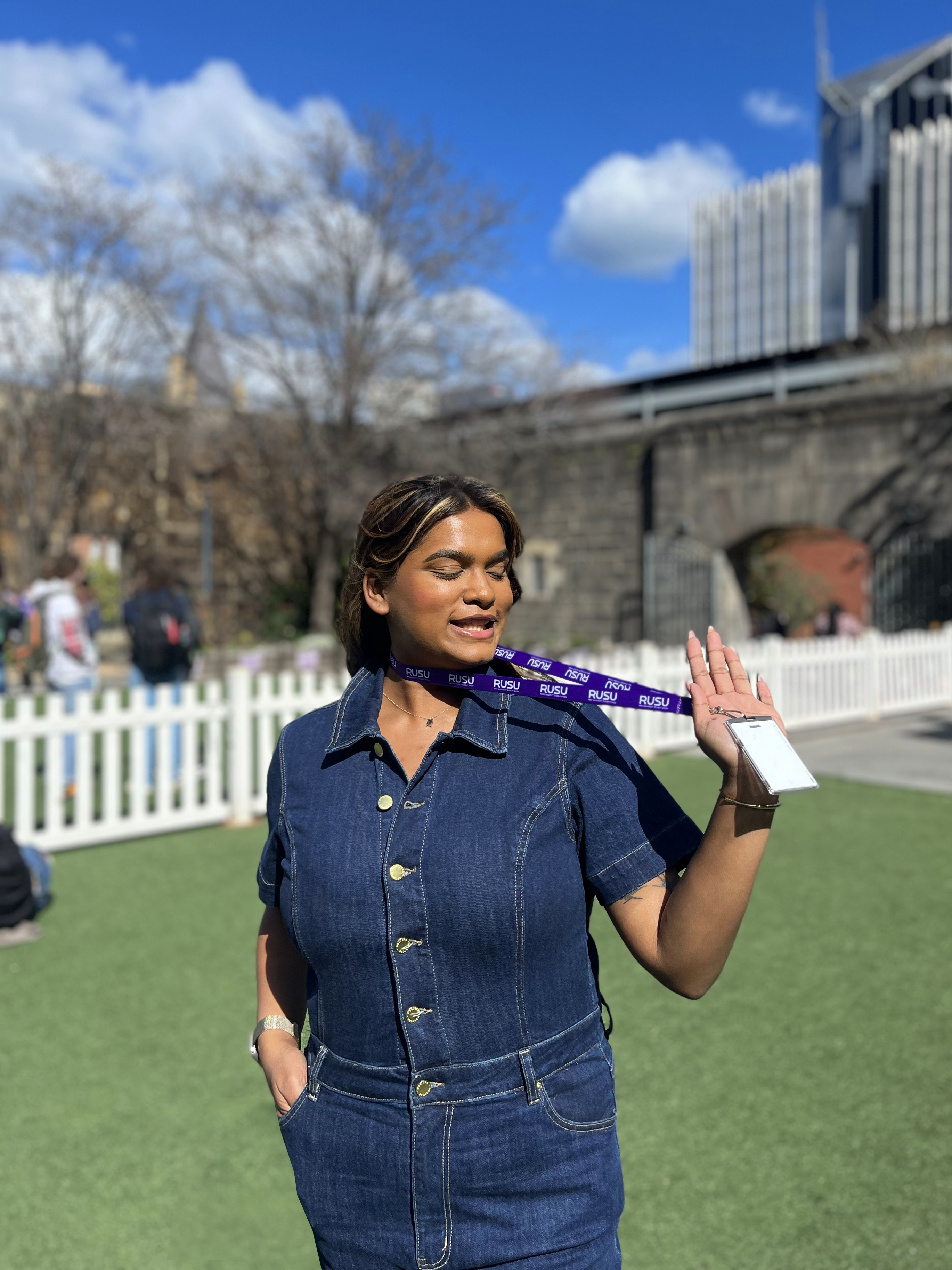 Model wearing a denim jumpsuit holds a purple RUSU lanyard from the RMIT Store, outdoors with buildings and blue sky in the background.