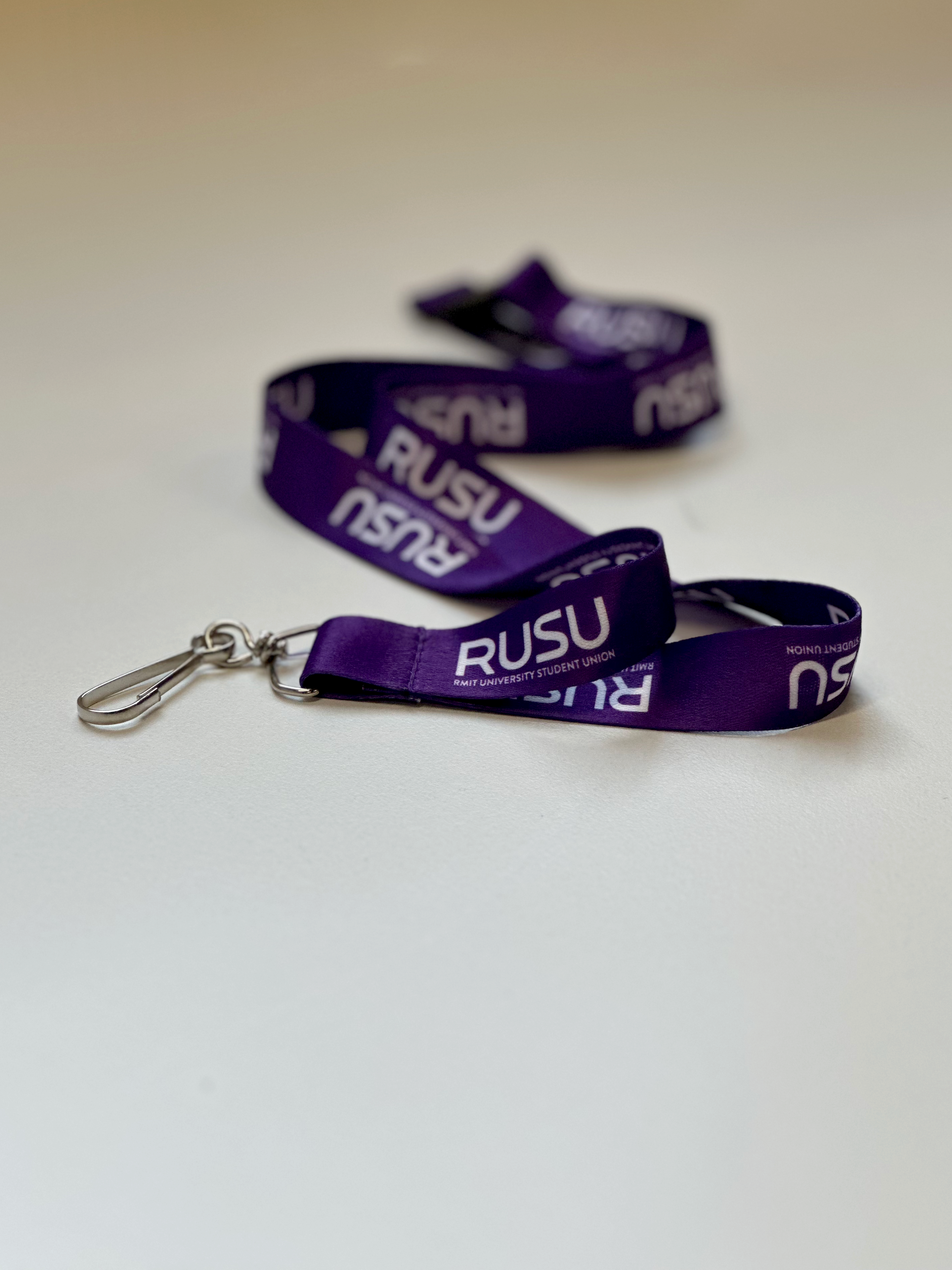 Close-up view of a purple RUSU lanyard with RMIT branding, highlighting the silver metal clip, resting on a white table.