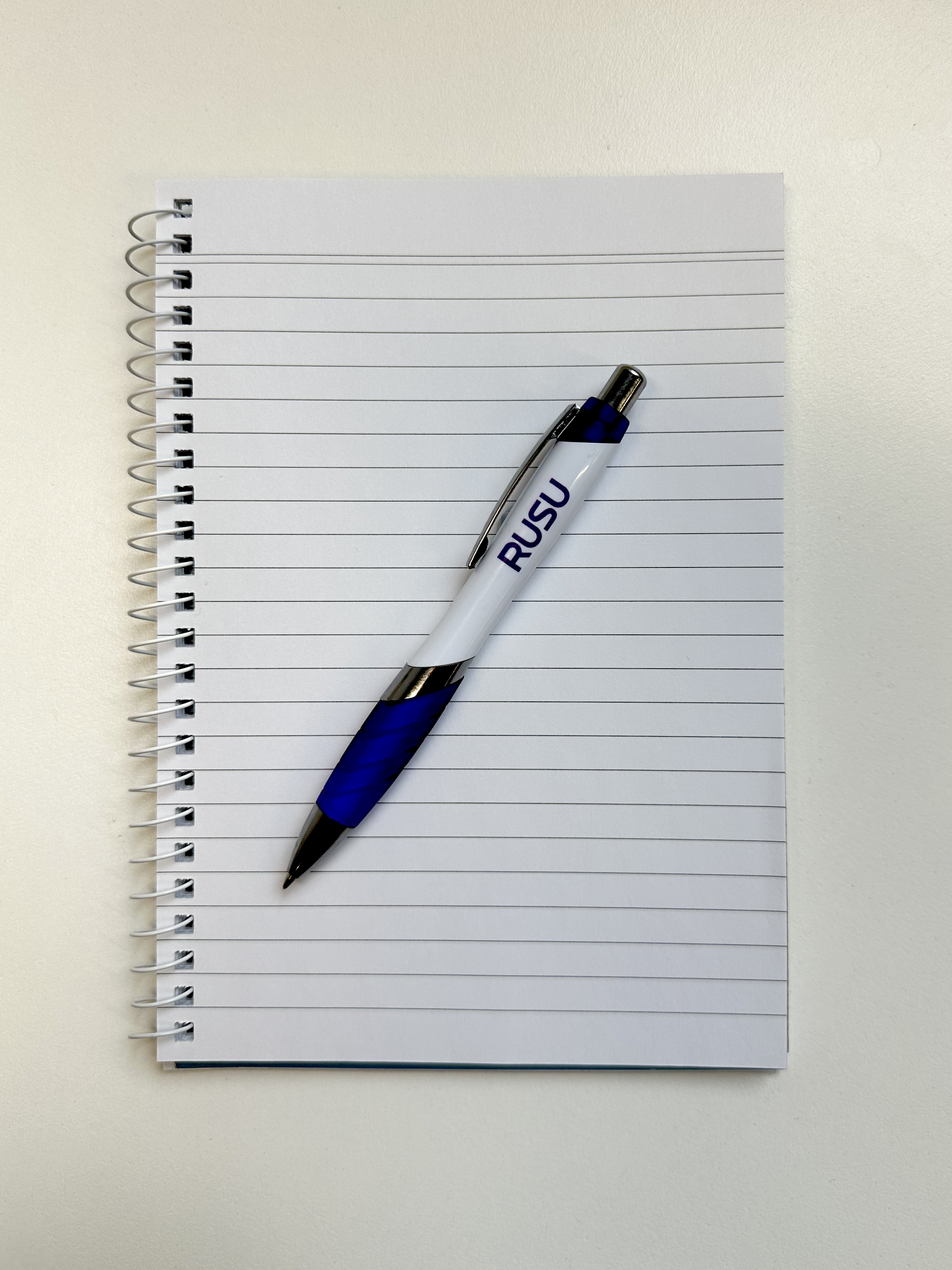 RUSU Pen in white and purple with a purple RUSU logo. Top down view lying flat on a notebook.
