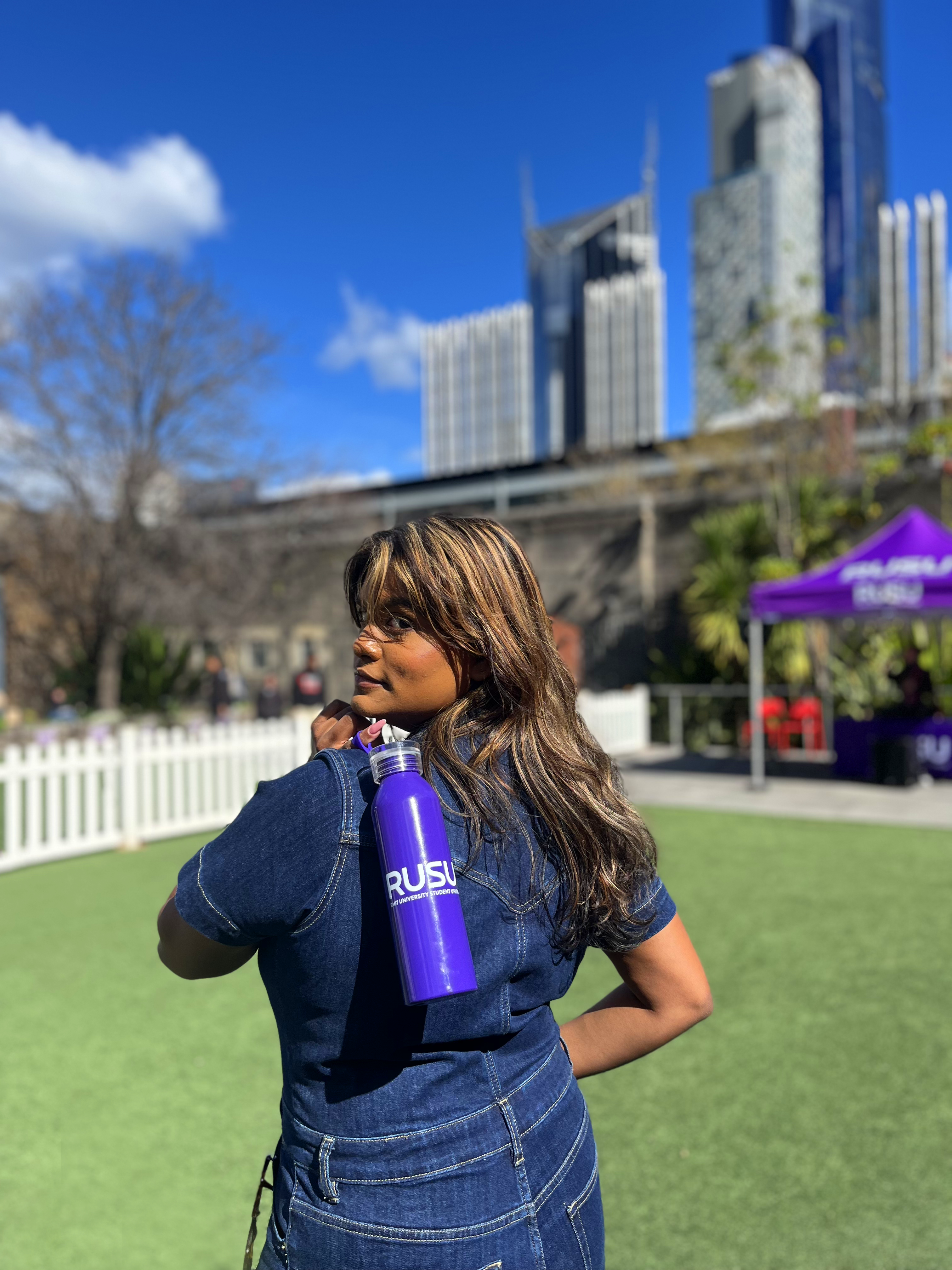 RUSU Water Bottle in puprple with a white printerd RUSU logo. Modelled being held by silicone strap with the bottle being helf over the shoulder with the photo taken from behind the model.