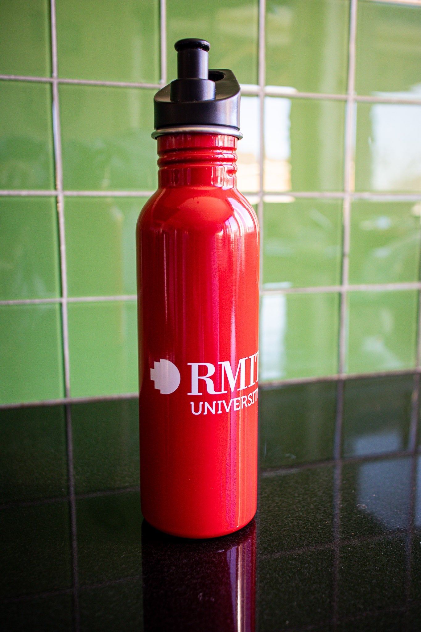Full view of the red RMIT bottle with a white RMIT University logo centered on the body, black cap securely on the bottle, placed on a black tabletop with a green background. Available at the RMIT Store.