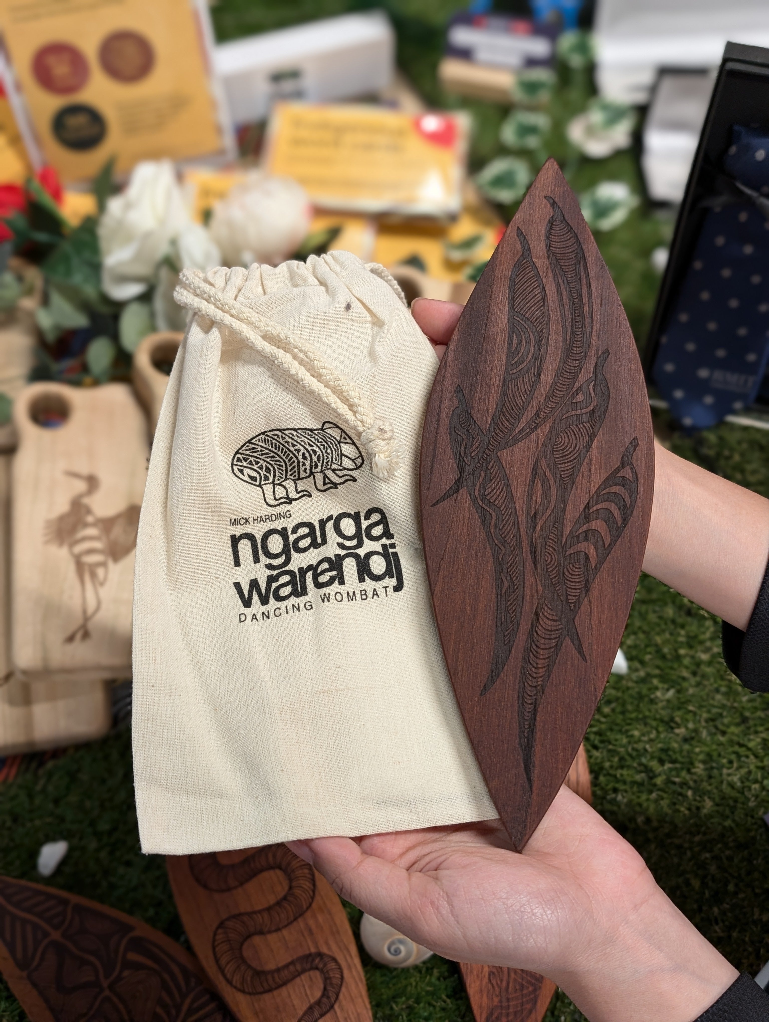 A hand holding a Red Cedar Malgarr Ngarga Warendj artefact with intricate Gum Leaves pattern carved into the dark wood, next to a beige Ngarga Warendj branded drawstring bag. The background features a variety of RMIT Store items, highlighting cultural craftsmanship.