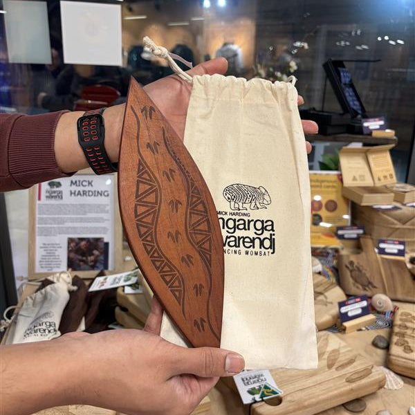 Model holding Red Cedar Malgarr – Ngarga Warendj Barring Tracks variant, featuring a curved, elongated red cedar wooden form with laser-etched geometric track patterns and repeated symbolic markings across the surface, paired with a natural cotton drawstring pouch printed with Mick Harding Ngarga Warendj Dancing Wombat logo and wombat illustration, displayed against an in-store RMIT Store wooden table setting with additional engraved boards and gift items in the background.