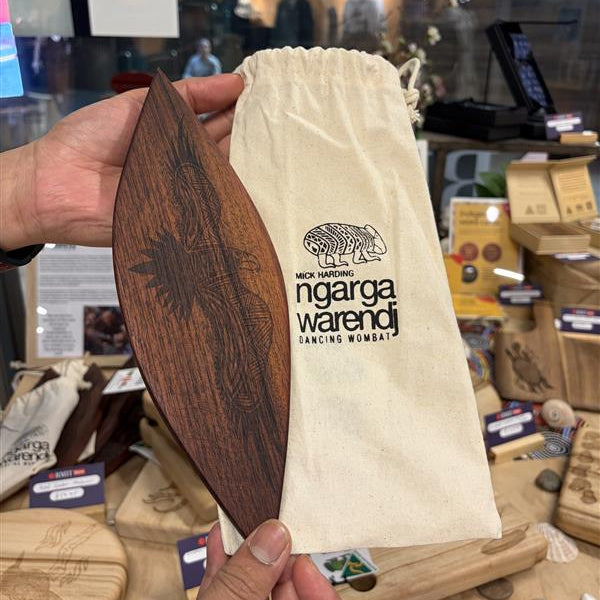 Model holding Red Cedar Malgarr – Ngarga Warendj Bunjil The Wedge Tailed Eagle variant, showing a dark red cedar carved piece with detailed laser-etched eagle-inspired linework and feather-like patterns across the surface, alongside a natural cotton drawstring pouch printed with Mick Harding Ngarga Warendj Dancing Wombat branding and wombat icon, photographed in-store at the RMIT Store with wooden serving boards and boxed merchandise visible in the background.