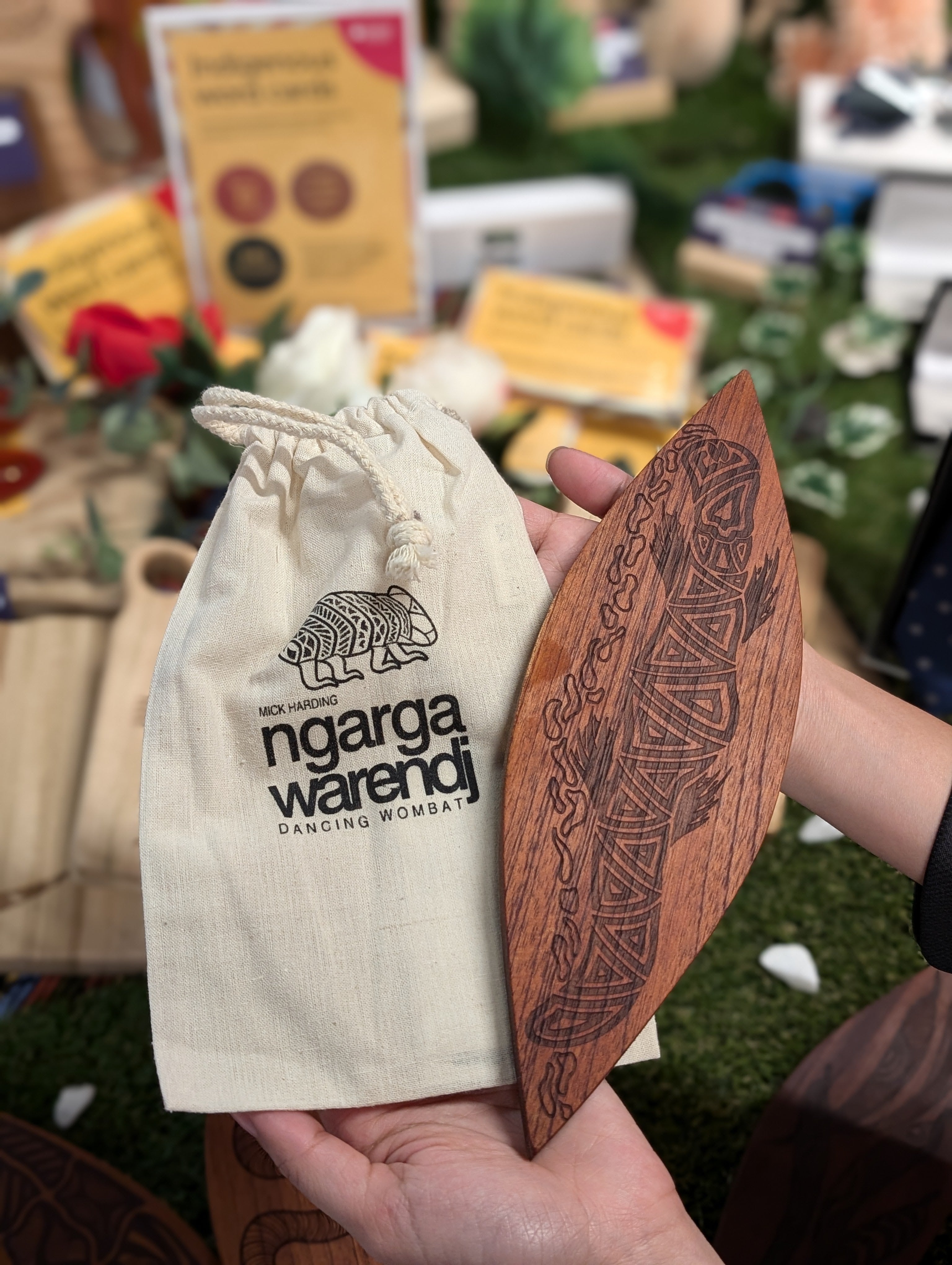 Hands displaying a Red Cedar Malgarr Ngarga Warendj artefact inscribed with branding and a Budja the Platypus design, featuring a mounting hook. The backdrop includes diverse Indigenous-inspired products, showcasing artisanal detail and cultural heritage.