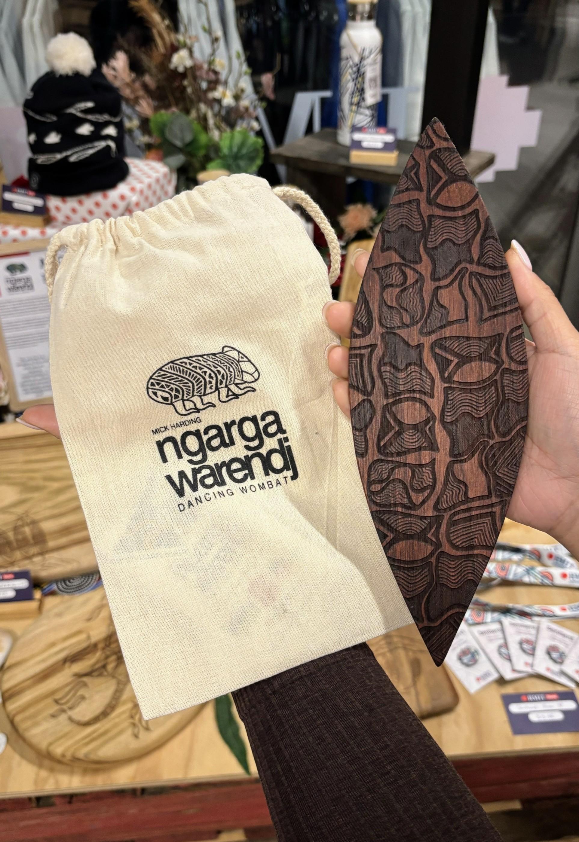 A Red Cedar Malgarr Ngarga Warendj artefact featuring a Gugra the Possum Skin Cloak pattern, held next to a Ngarga Warendj branded bag. The setting is filled with indigenous items, underscoring the artefact's artistic expression and traditional roots.