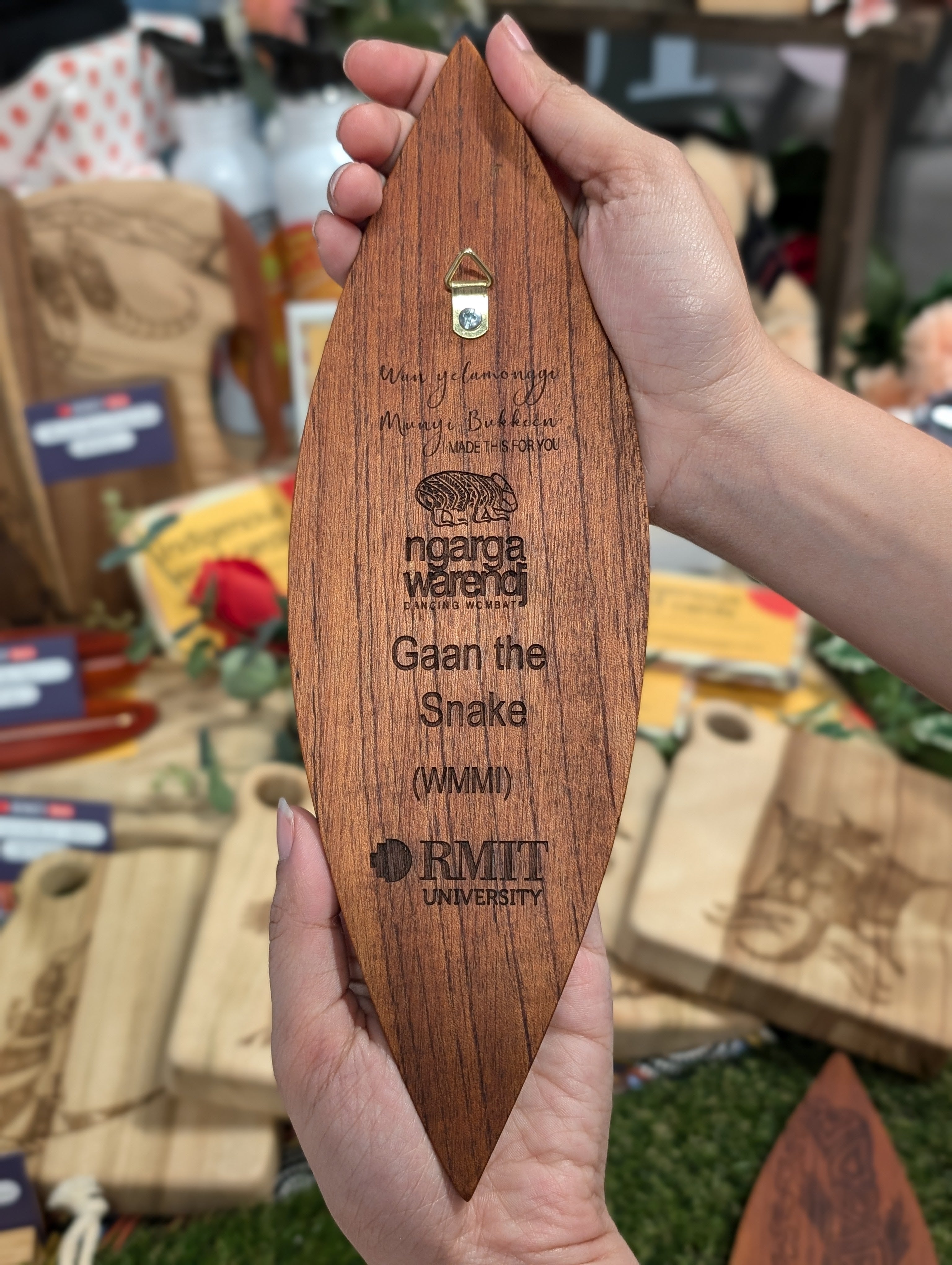 Rear view of a Red Cedar Malgarr Ngarga Warendj artefact displaying engraved branding and a Gaan the Snake inscription, featuring a mounting hook. The backdrop includes diverse Indigenous-inspired products, showcasing artisanal detail and cultural heritage.