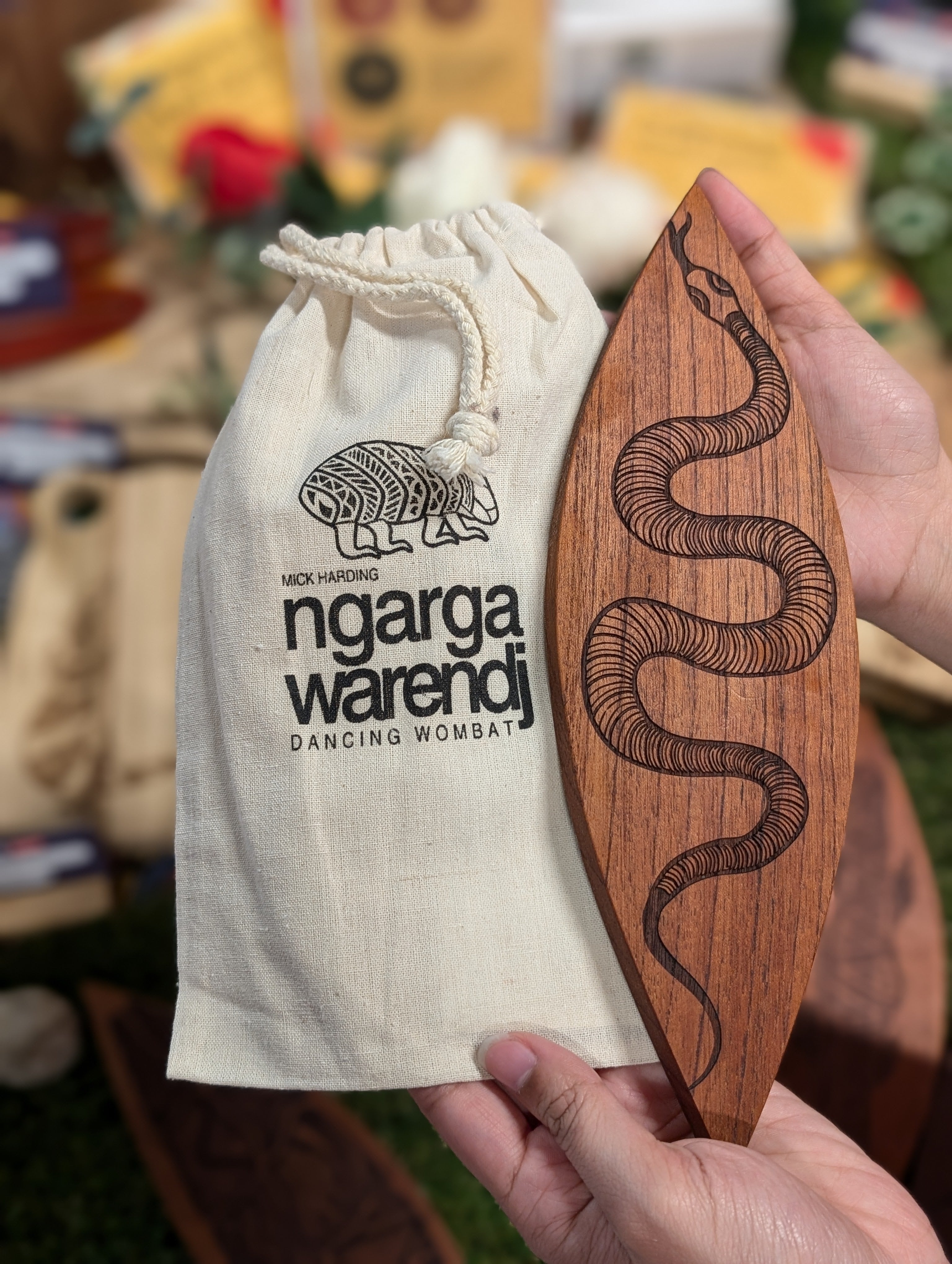 Two hands presenting a Red Cedar Malgarr Ngarga Warendj artefact with an engraved Gaan the Snake design, accompanied by a Ngarga Warendj branded bag. The surrounding colourful flowers and Indigenous word cards accentuate the item's cultural significance.