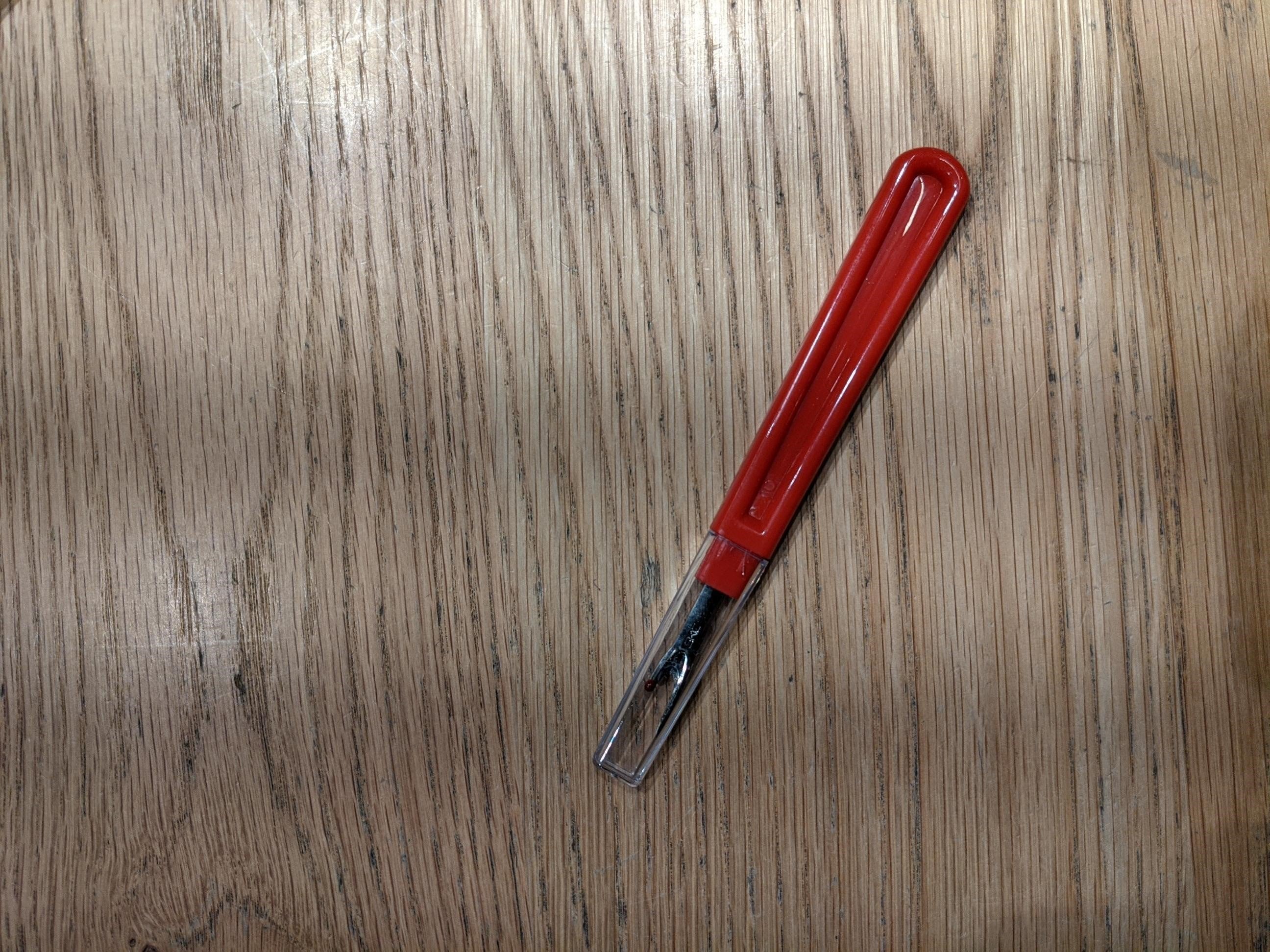 Red seam unpicker with a curved plastic handle and clear protective cap, shown laid horizontally on a beige fabric tabletop. The metal blade features a red safety ball on the shorter tip. Available at the RMIT Store.