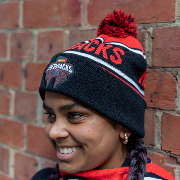 Model wearing a Redbacks beanie in red and black, highlighted by bold red lettering 'REDBACKS' and an embroidered logo patch near the forehead, showcasing the Redbacks emblem. The beanie features a red pom-pom and striped detailing in white, red, and black. The model wears a matching scarf, braided hair, and small gold hoop earrings, posed against a textured brick wall. Part of the RMIT Store collection.