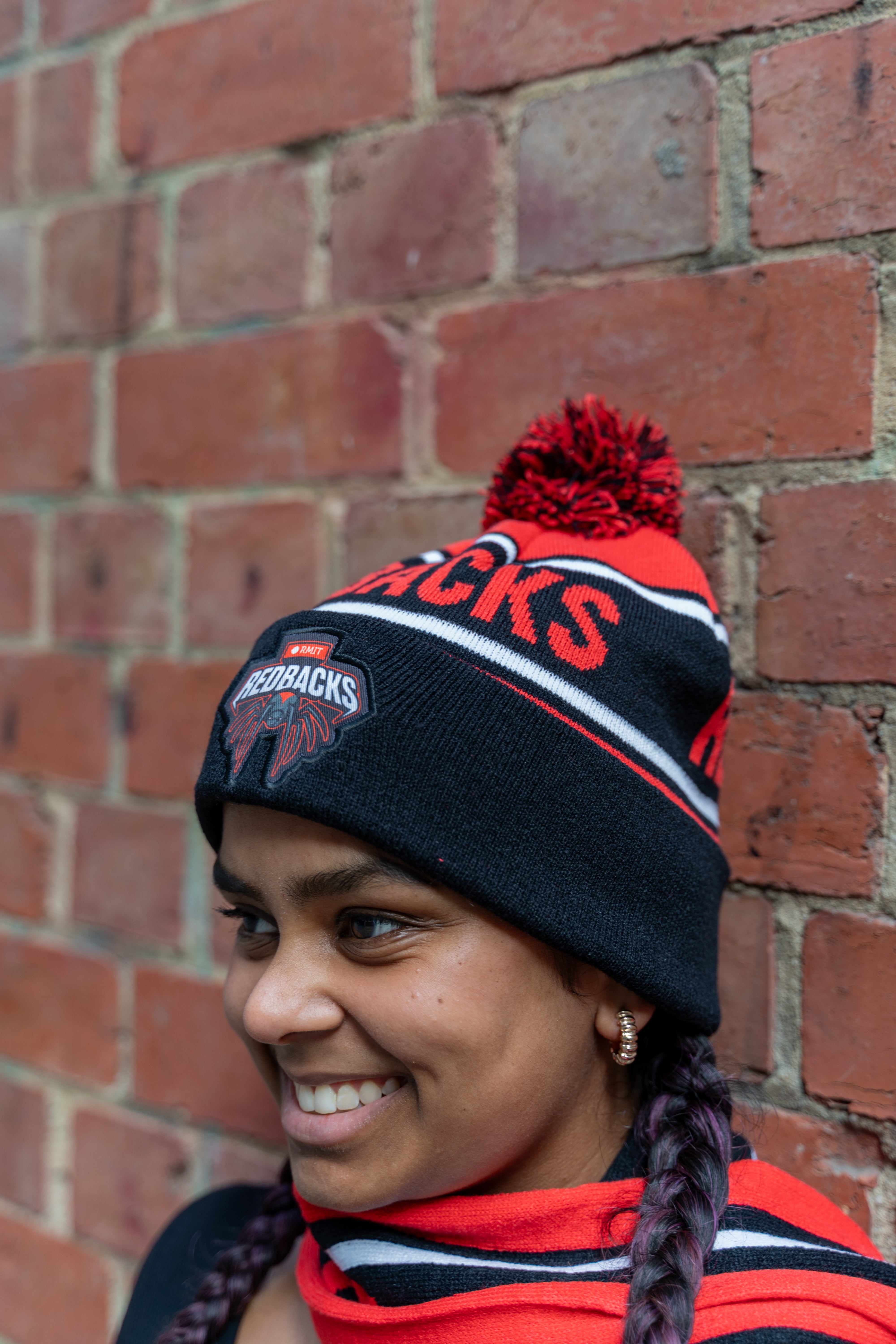 Model wearing a Redbacks beanie in red and black, highlighted by bold red lettering 'REDBACKS' and an embroidered logo patch near the forehead, showcasing the Redbacks emblem. The beanie features a red pom-pom and striped detailing in white, red, and black. The model wears a matching scarf, braided hair, and small gold hoop earrings, posed against a textured brick wall. Part of the RMIT Store collection.