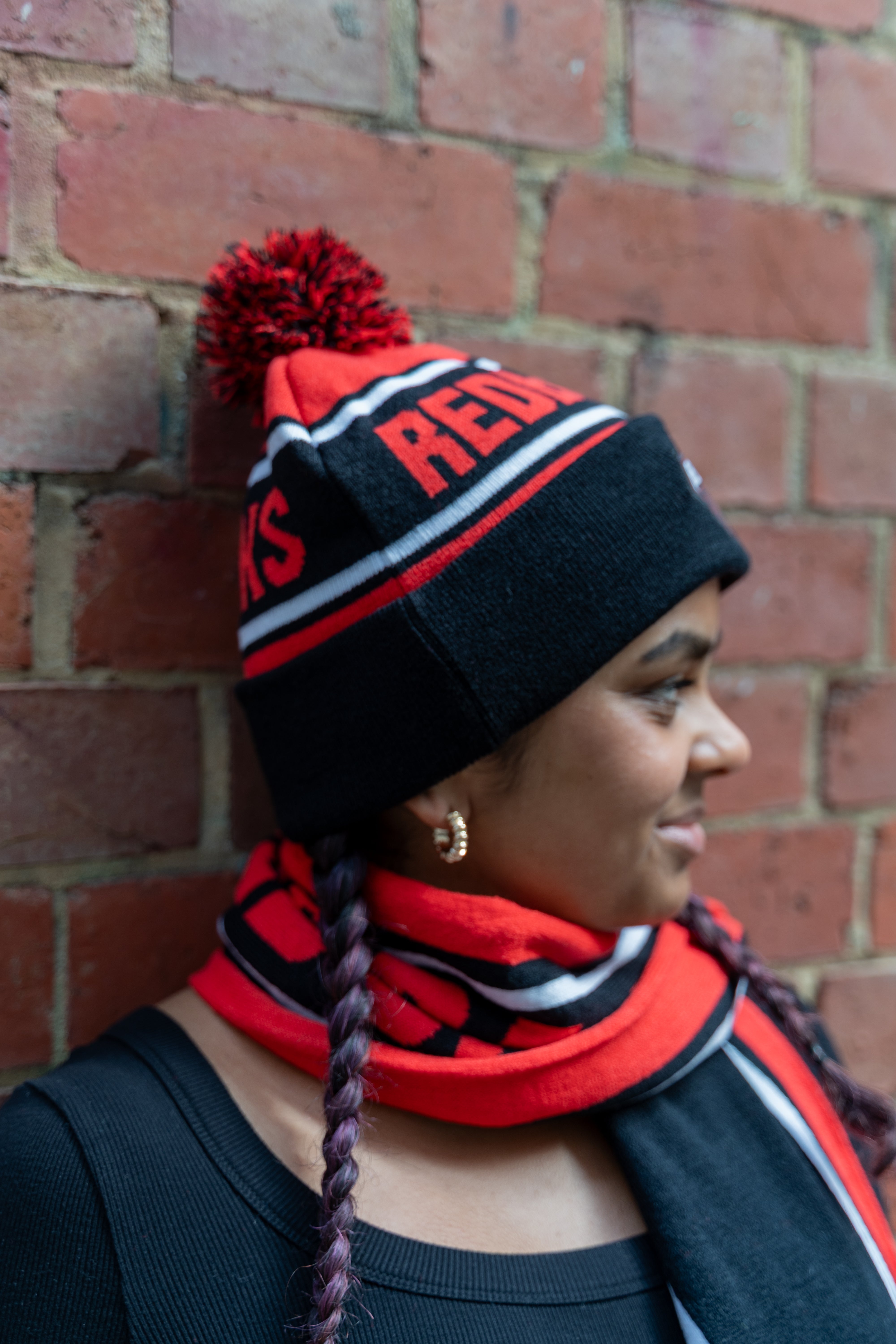 Model wearing a Redbacks beanie in red and black, featuring bold red lettering 'REDBACKS' on a black band. The beanie includes a red pom-pom on top and subtle white, red, and black stripe details. The model has braided hair and wears gold hoop earrings, with a matching red and black Redbacks Scarf. Visible in an urban setting against a brick wall. Part of the RMIT Store collection.