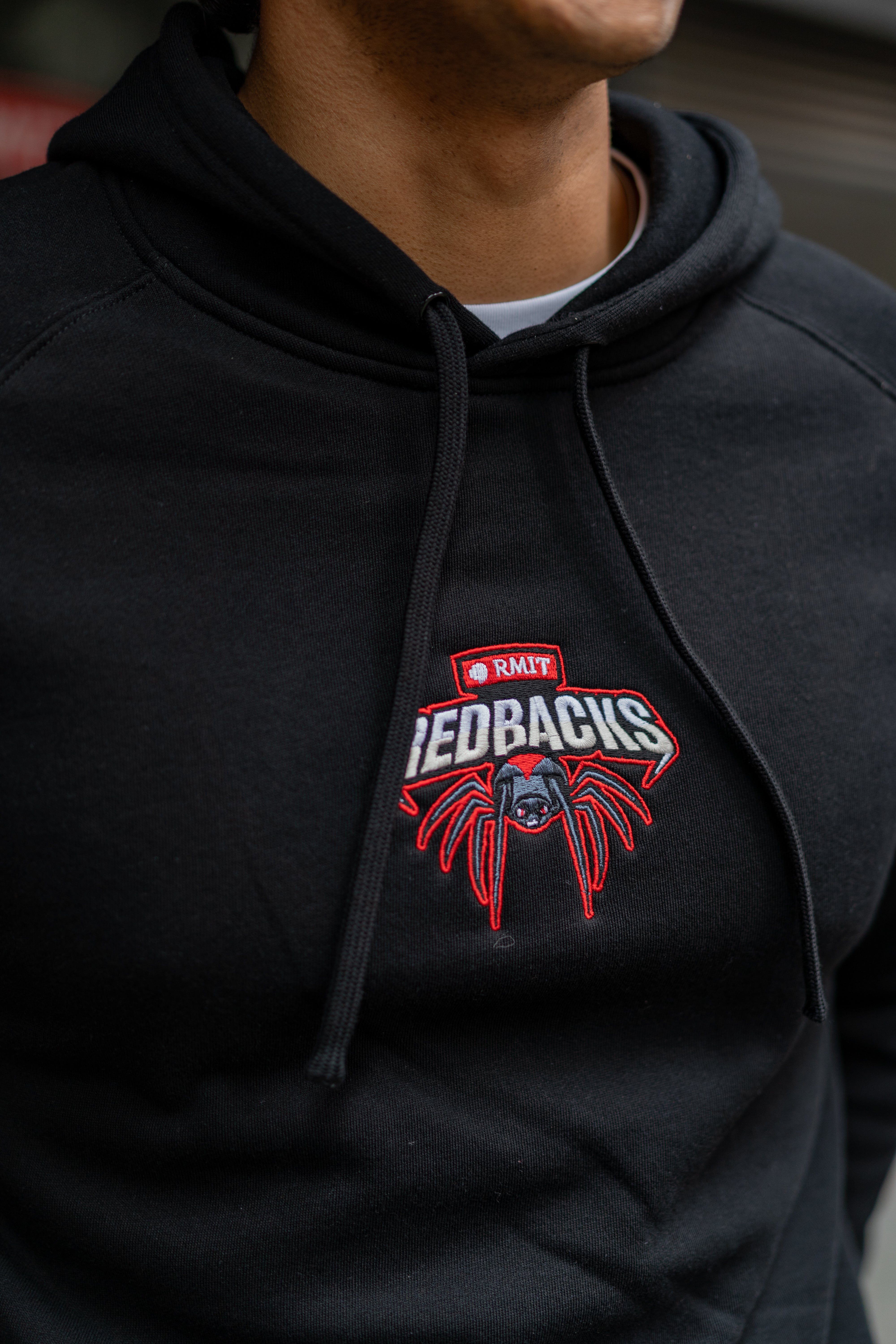 Close-up detail of a black Redbacks Hoodie worn by a male model, featuring embroidered red and white RMIT Redbacks spider logo on the chest, black drawstrings and soft fleece fabric detail, available at the RMIT Store.