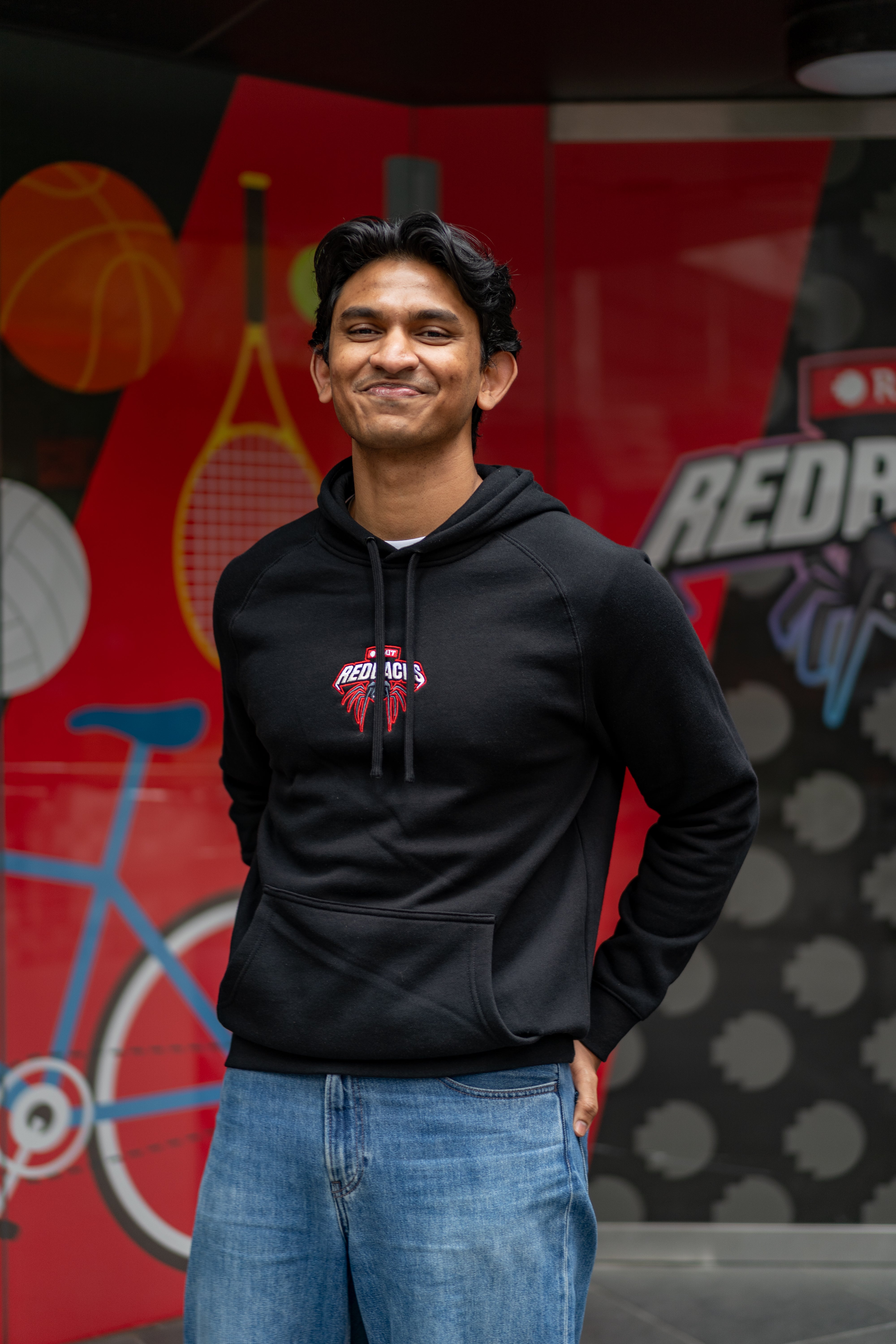 Male model wearing a black Redbacks Hoodie with embroidered red and white Redbacks spider logo on the chest, black drawstrings and kangaroo pocket, standing in front of a sports-themed mural featuring bicycle and tennis graphics, available at the RMIT Store.