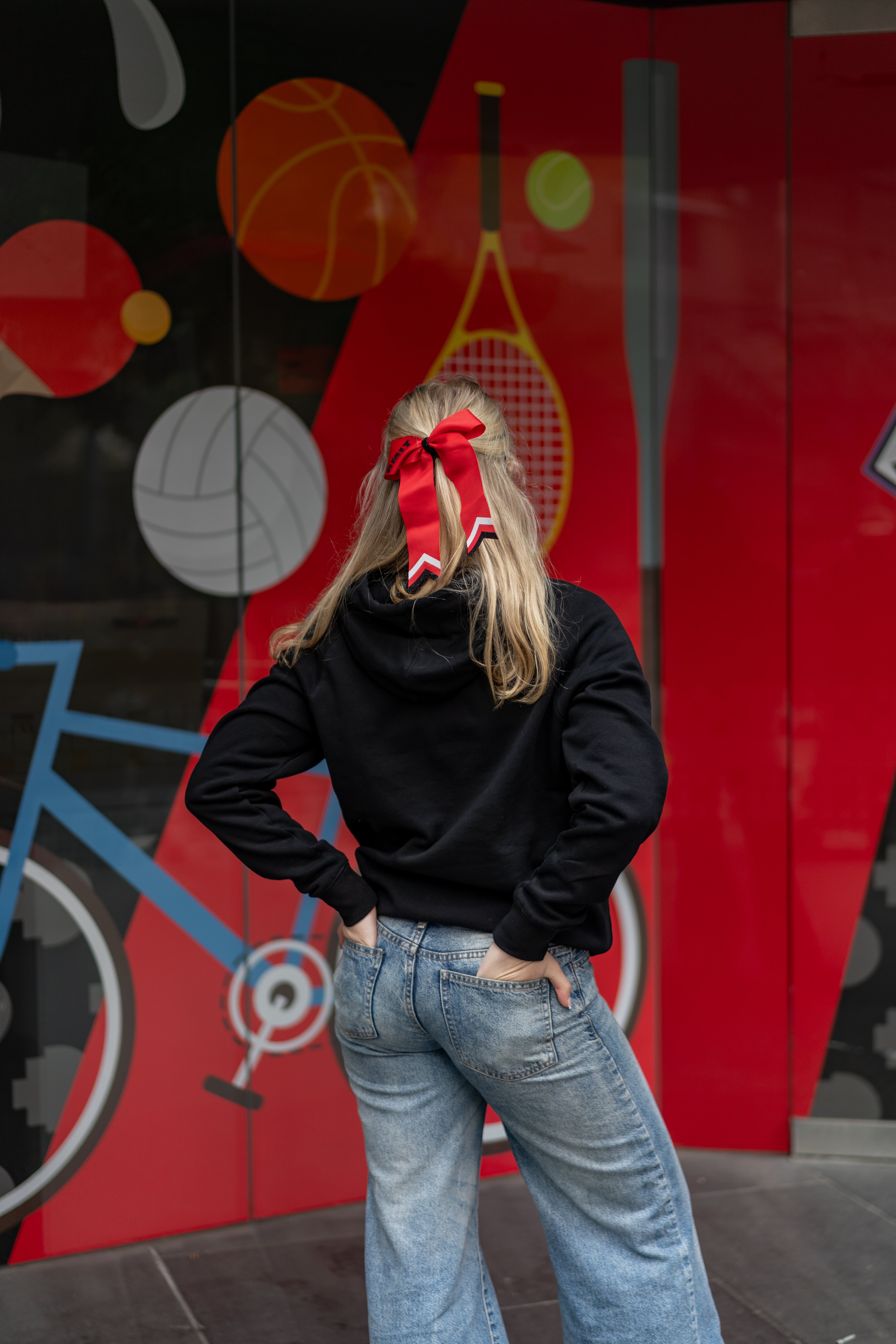 Rear view of a female model wearing a black Redbacks Hoodie with hood detail and ribbed waistband, paired with a large red bow in blonde hair, standing in front of a colourful sports-themed wall with tennis and cycling illustrations, available at the RMIT Store.