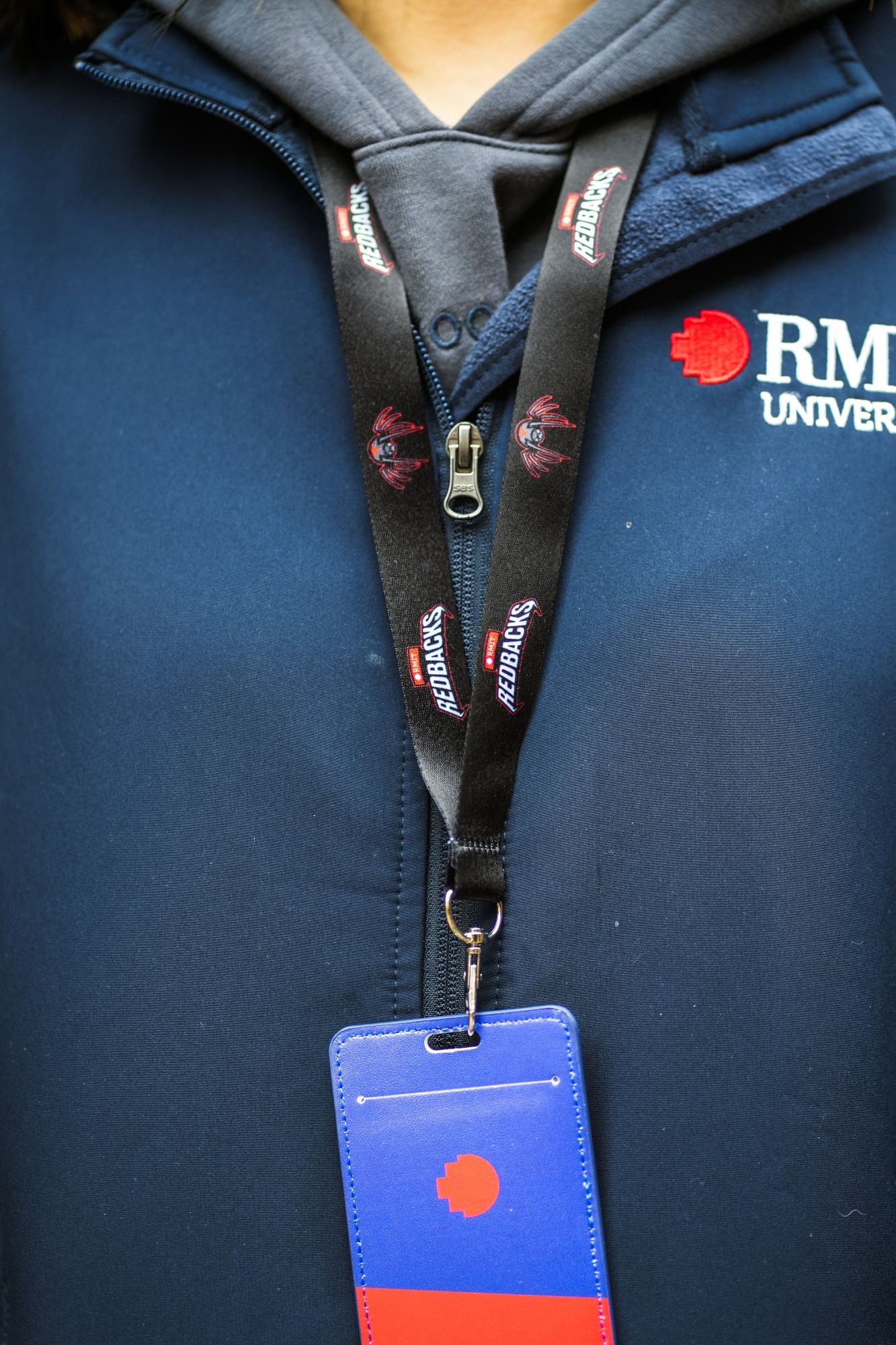 A model wearing a navy blue RMIT softshell jacket showcases a black Redbacks lanyard adorned with the team's logo and mascot in red and white. The lanyard, hanging around the neck, supports a blue and red RMIT card holder at chest level. This stylish lanyard is part of the selection available at the RMIT Store.