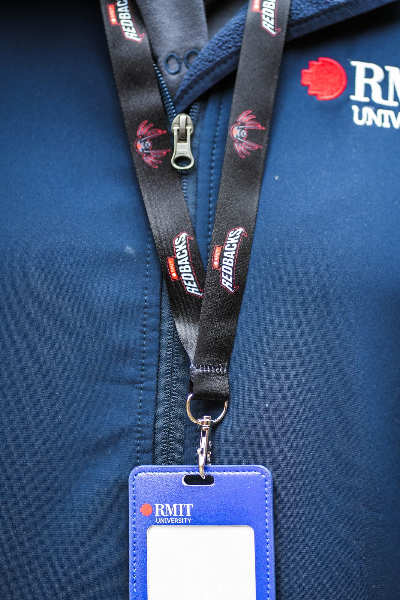 A black Redbacks lanyard with the team's branding and mascot visible in red and white is worn by a model on a navy RMIT softshell jacket. The lanyard supports a blue and red card holder, prominently displaying the university logo, highlighting a practical and stylish accessory found in the RMIT Store.