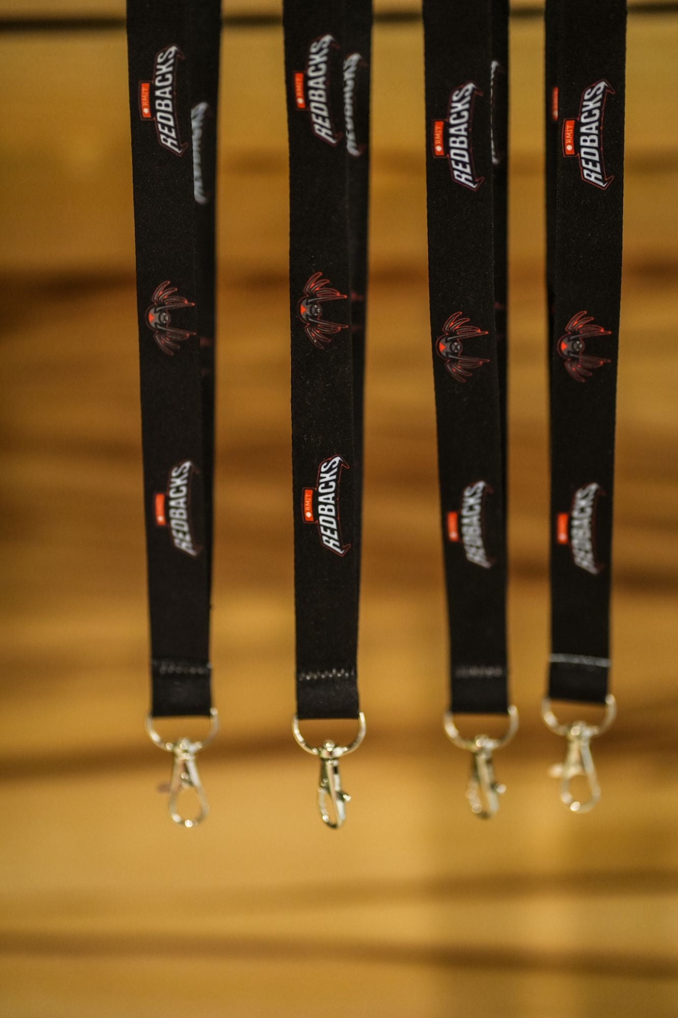 Four black Redbacks lanyards are hanging against a wooden background, showing the team's logo and mascot in a repeated pattern. Each lanyard features a silver swivel hook at the end, accentuating the vibrant red and white branding against the dark fabric. This close-up highlights the sleek design offered in the RMIT Store.