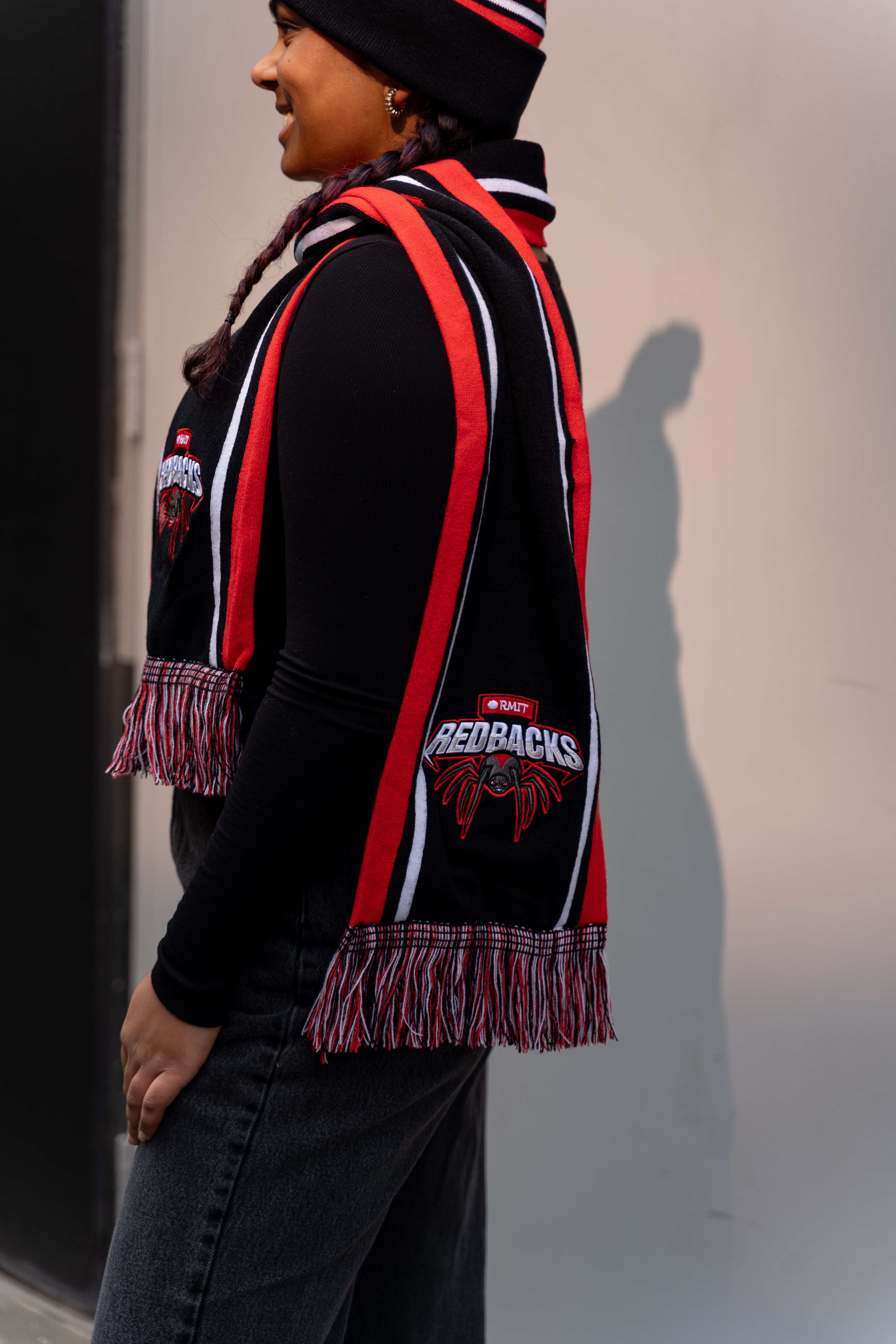 Model wearing the Redbacks scarf with bold red, black, and white stripes, featuring an embroidered Redbacks logo and spider design along with the RMIT branding. The scarf has multicoloured fringes at the ends, and is worn over a black top in a side studio setting. Highlighting the scarf's distinct RMIT Store branding detail.