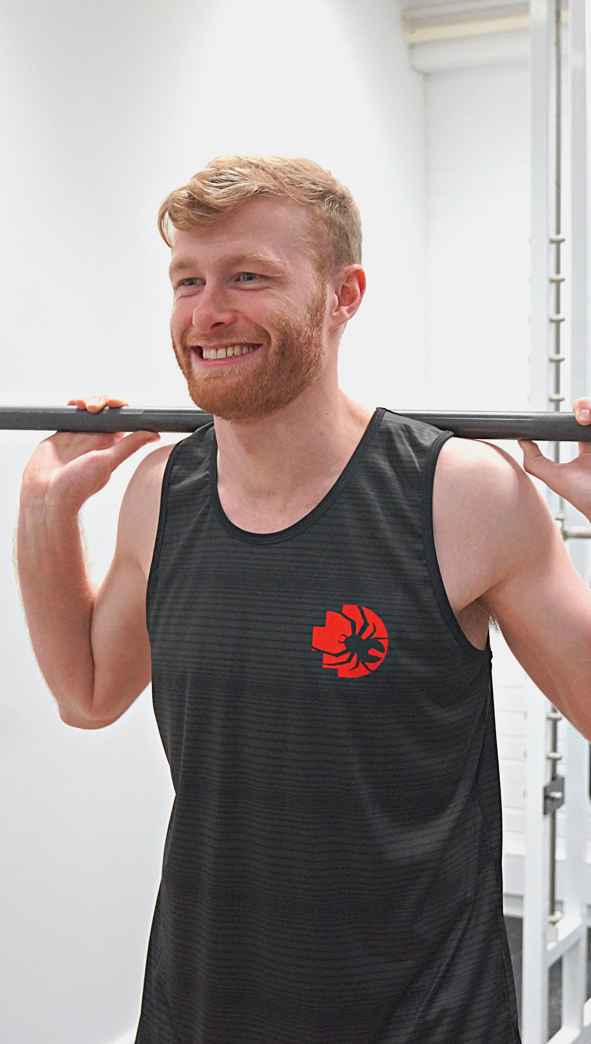 Male model wearing the RMIT Redbacks Singlet in black with red and black Redbacks logo on the left chest, smiling while lifting weights. The singlet has subtle light black horizontal patterns. The model is indoors in a gym setting. Available at the RMIT Store.