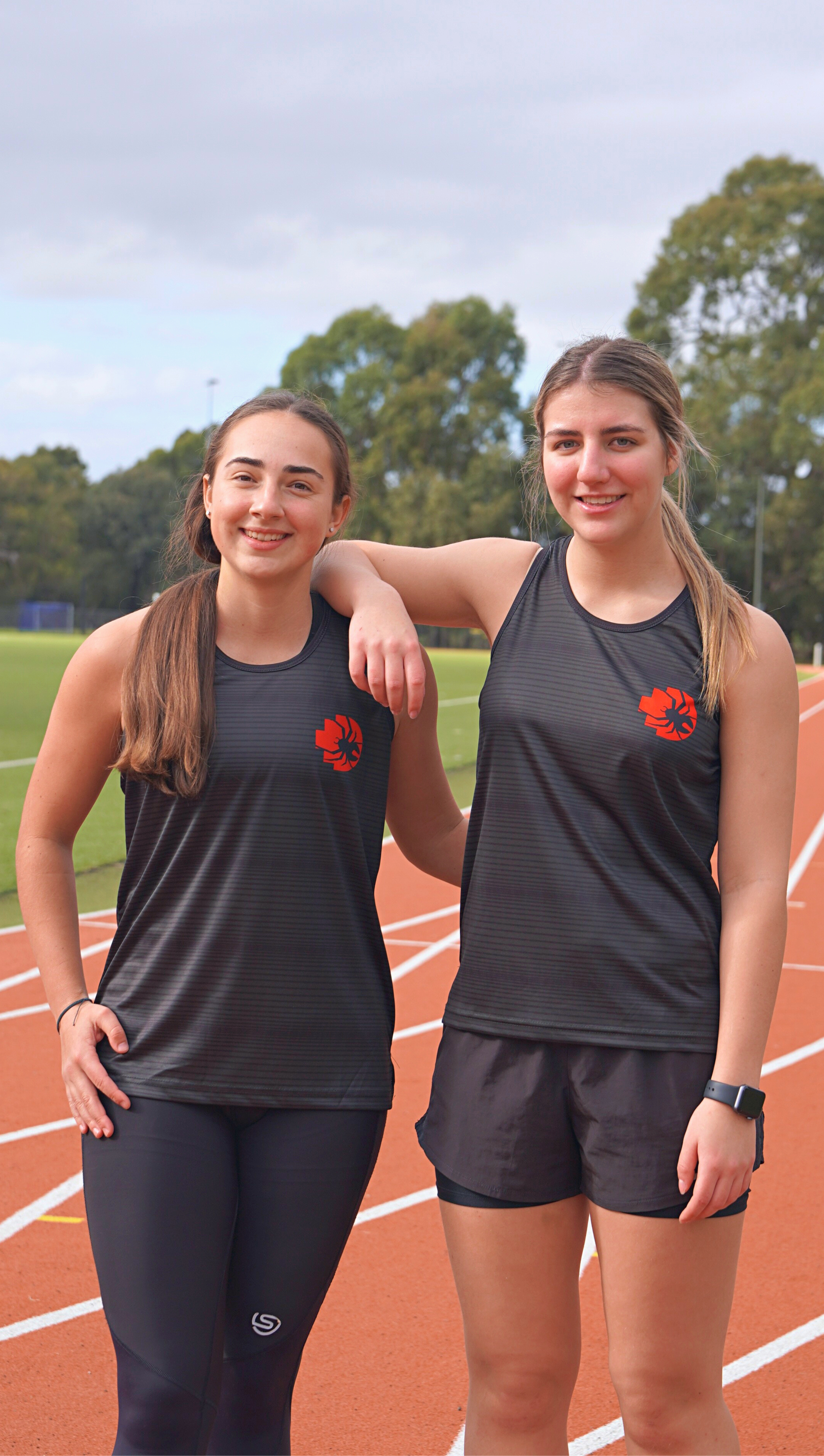 Two female models standing front view on running tracks wearing black RMIT Redbacks Singlets with red and black Redbacks logos on left chests, the model on the left resting her right arm on the right model’s shoulder, both smiling, subtle light black horizontal patterns visible on singlets. Available at the RMIT Store.
