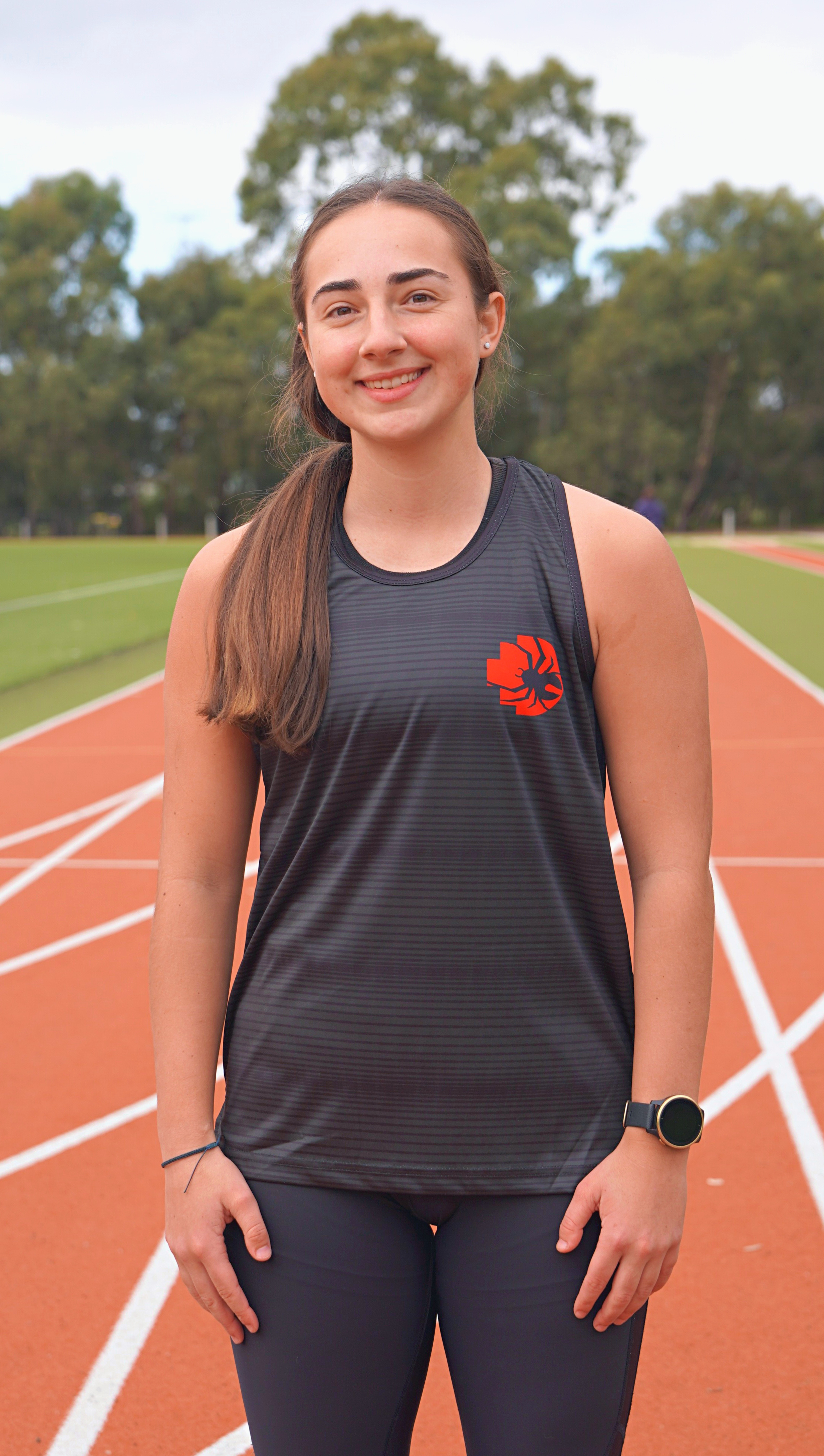 Female model smiling and wearing the RMIT Redbacks Singlet in black with red and black Redbacks logo on the left chest, standing outdoors on running tracks. The singlet features subtle light black horizontal patterns. Available at the RMIT Store.
