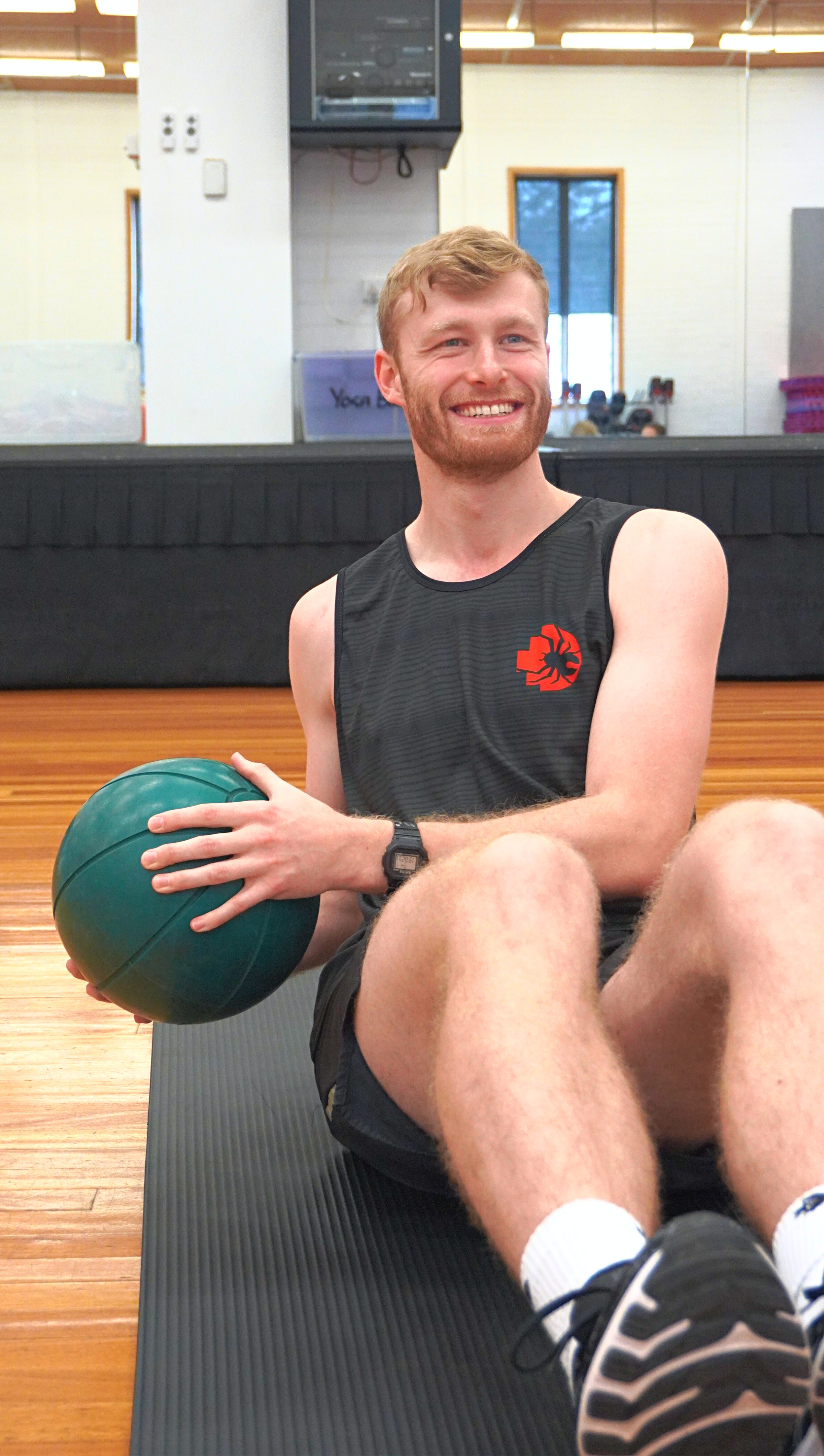 Male model wearing the RMIT Redbacks Singlet in black with red and black Redbacks logo on the left chest, sitting on a gym floor mat holding a ball to his left side with both hands, gym environment visible. The singlet has subtle light black horizontal patterns. Available at the RMIT Store.