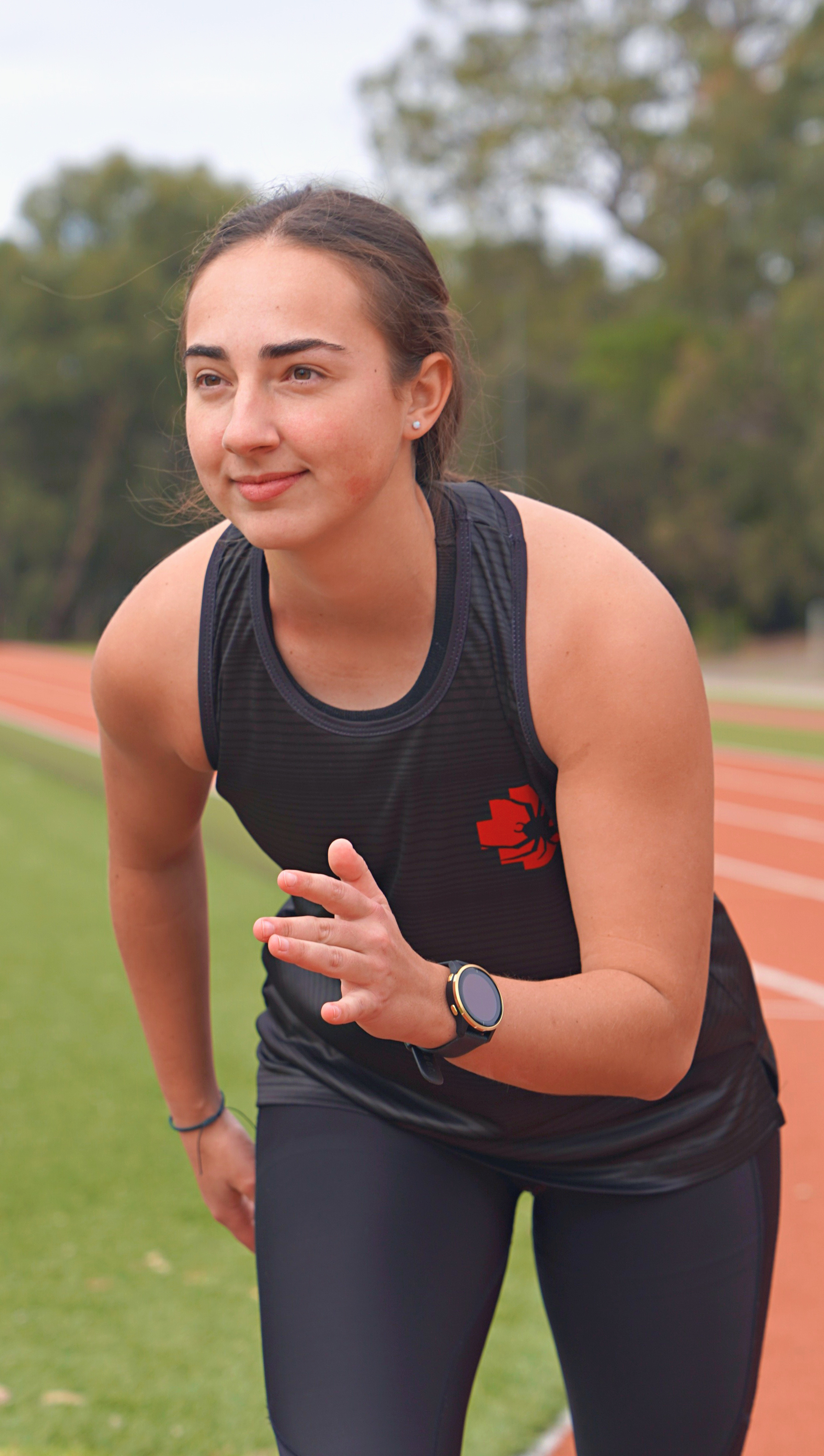 RMIT Redbacks Singlet in black worn by a female model posing in a starting position on an outdoor running track. The singlet features a red and black Redbacks logo on the left chest. Model is in a dynamic stance preparing to race, with visible sports lines behind her. Available at the RMIT Store.
