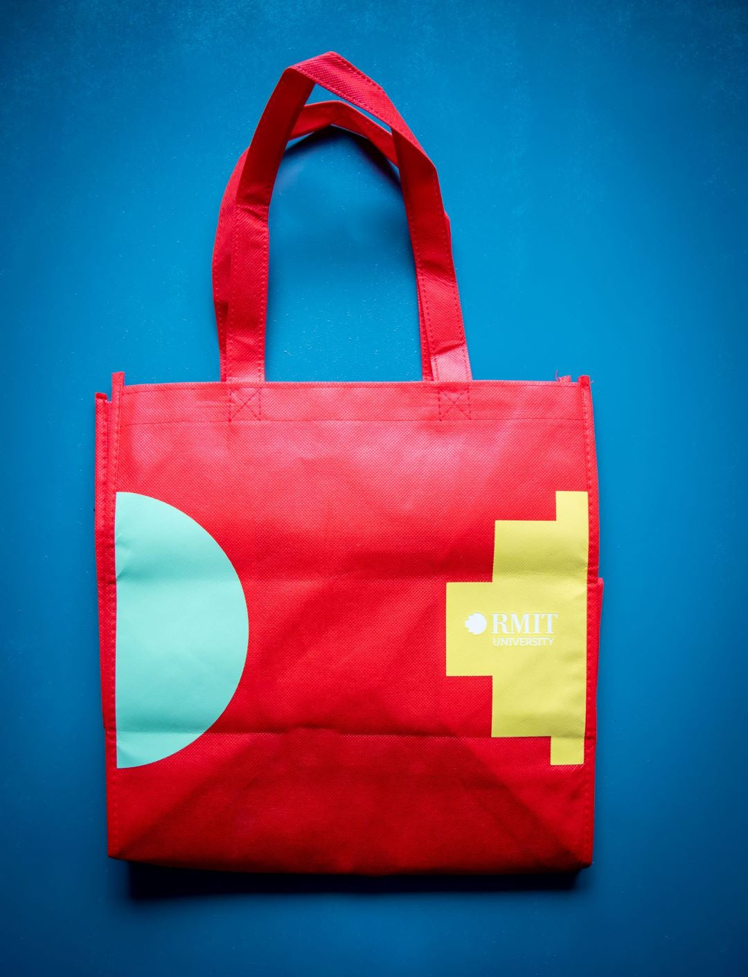 Bright red RMIT Reusable Bag with RMIT University logo in yellow and teal geometric design from the RMIT Store. The durable tote features dual handles, perfect for keeping course materials organised, displayed on a vibrant blue background.