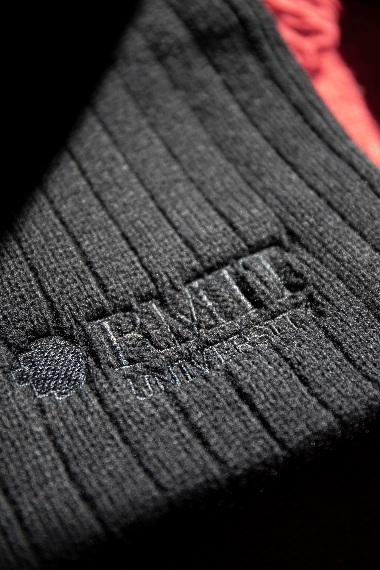 Close-up of the RMIT Ribbed Scarf showcasing its detailed ribbed texture and embossed RMIT University logo. The black fabric is contrasted against a vibrant red background in the sunlight, highlighting the scarf's intricate design.