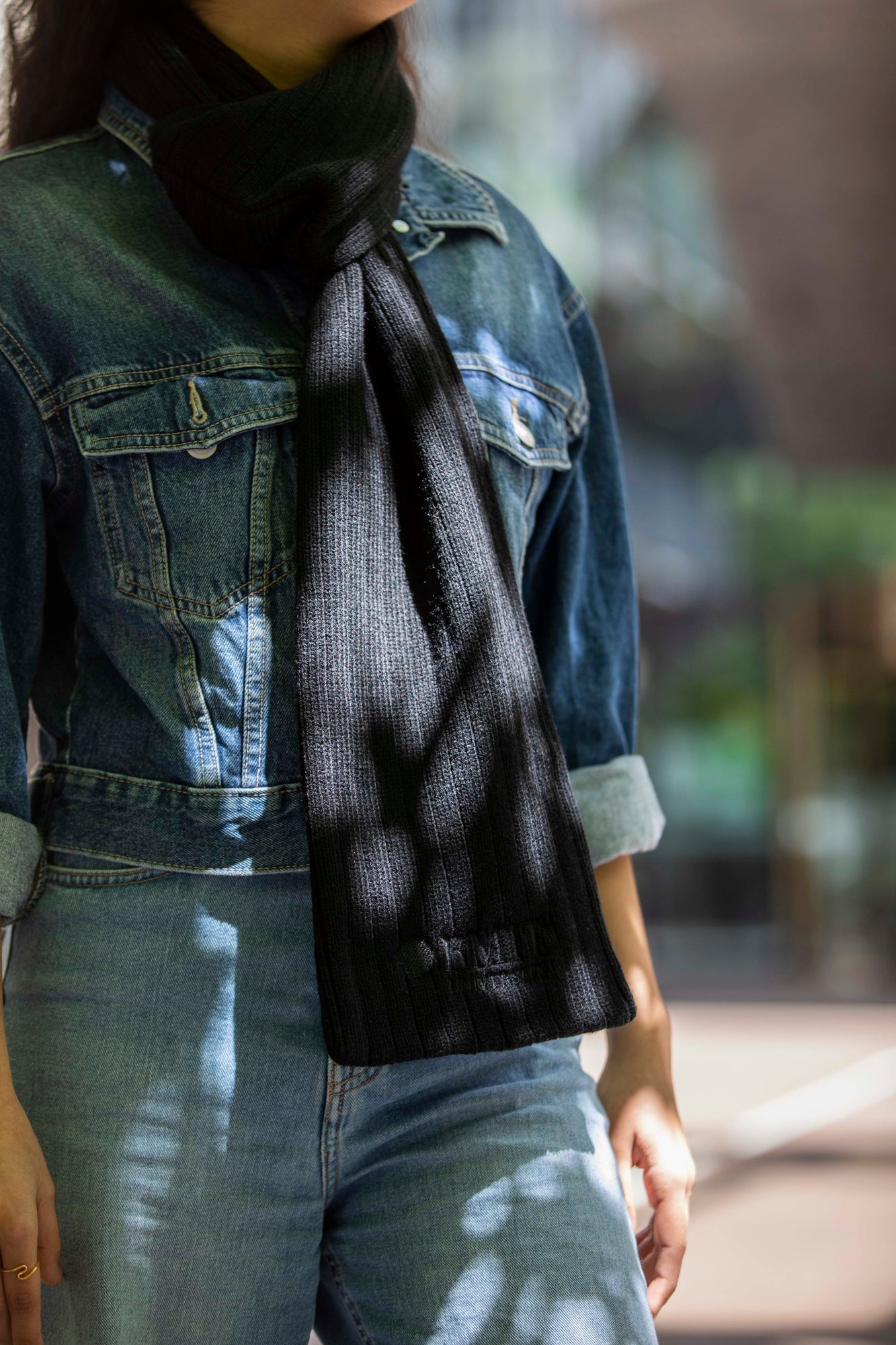 A model wearing the RMIT Ribbed Scarf, styled with a denim jacket and jeans. The black ribbed scarf is elegantly wrapped around the neck, its texture and warmth highlighted by the soft sunlight filtering through leaves.