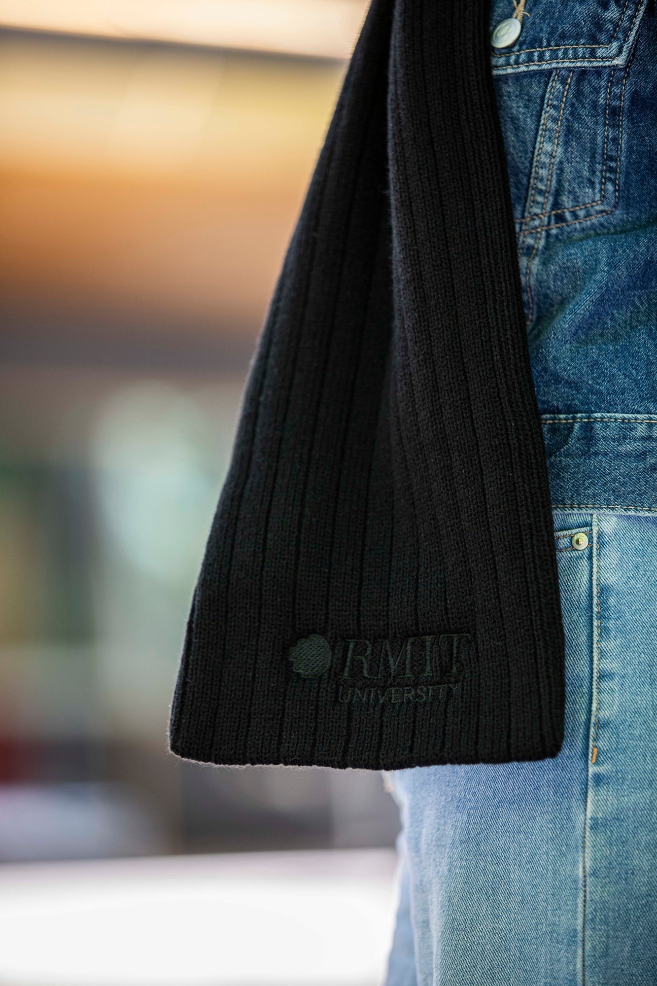 Side view of the RMIT Ribbed Scarf worn by a model in denim attire. The scarf's subtle ribbed pattern is accentuated, and the RMIT logo is visible towards the scarf's end. Soft, natural lighting adds depth to the textured fabric.
