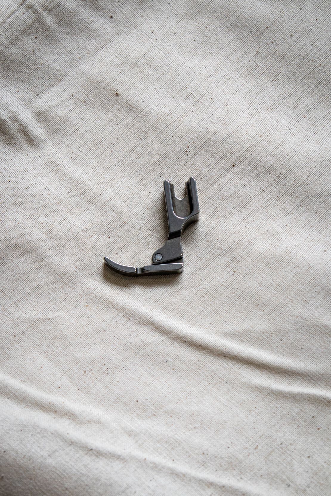 Angled side view of a silver right zipper foot sewing machine attachment placed on a pale beige surface, available from the RMIT Store.