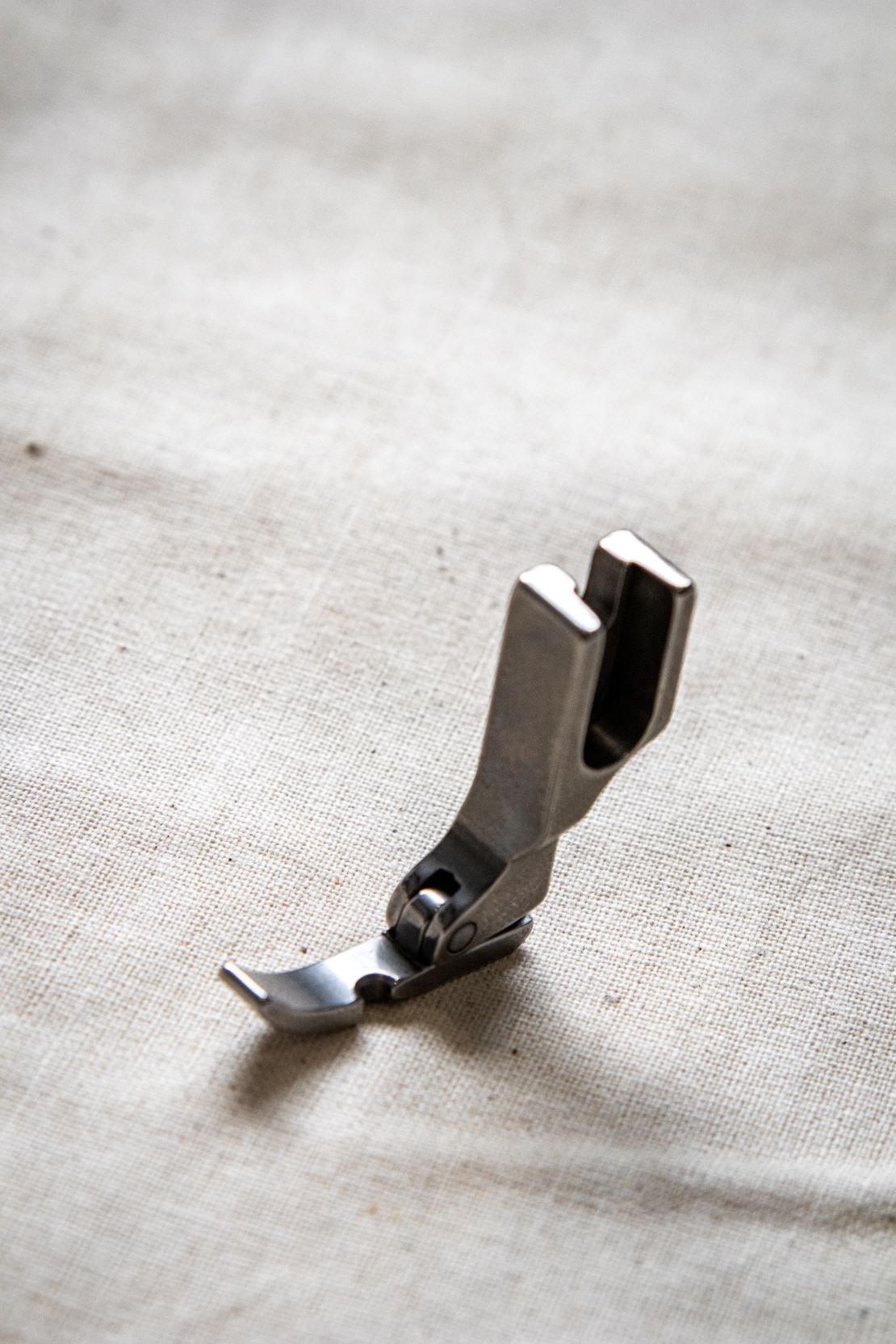 Close up side view of a silver right zipper foot sewing machine attachment on a pale beige surface, showing a narrow metal shape with a cut-out channel. Available from the RMIT Store.