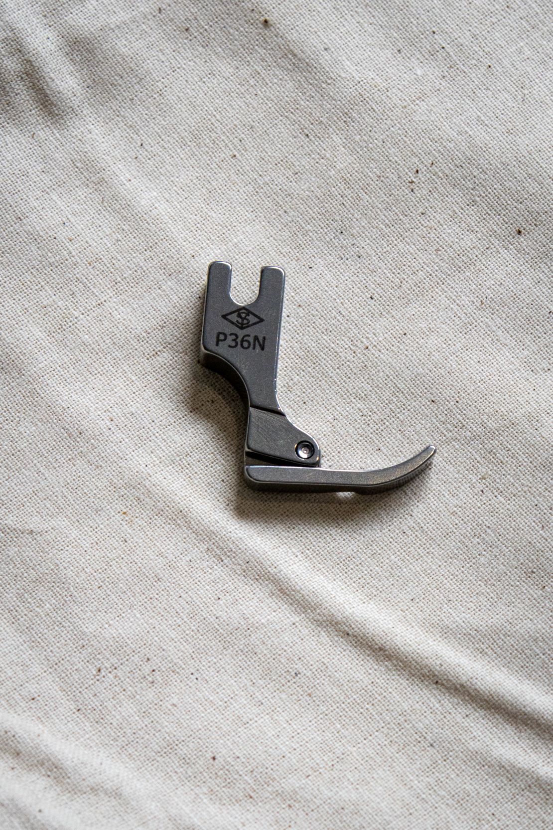 Close-up top view of a silver right zipper foot sewing machine attachment placed on a pale beige background, showing a clear engraving "P36N". Available from the RMIT Store.