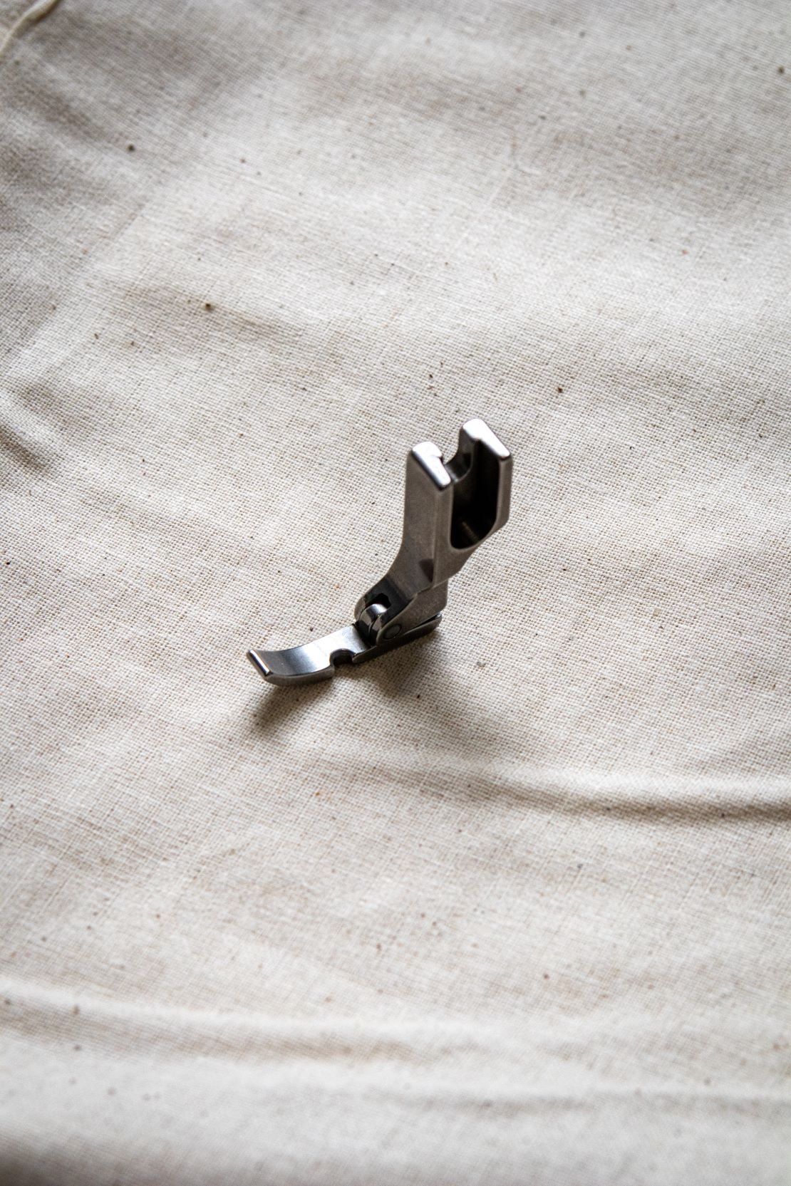 Side view of a silver right zipper foot sewing machine attachment on a pale beige surface, showing a narrow metal shape with a cut-out channel. Available from the RMIT Store.