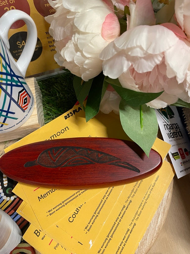 Rosewood Oval Pen Set featuring a polished reddish-brown oval wooden pen case with black laser-engraved geometric leaf pattern and fine line detailing, displayed on stacked yellow information cards beside artificial pink-and-white flowers and a patterned ceramic mug at RMIT Store, with no model present and subtle wood grain visible under the engraving.