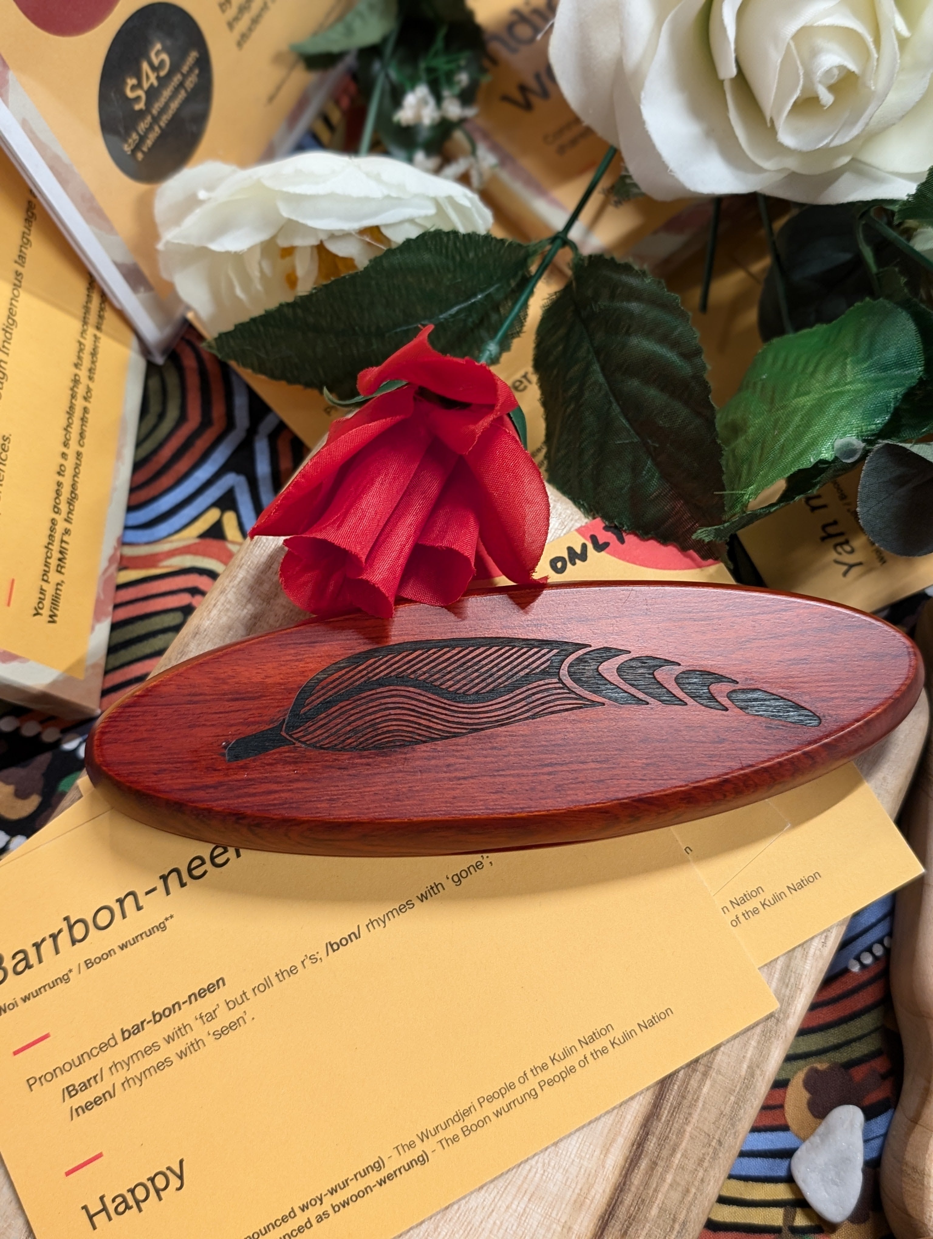 A rosewood pen set from the RMIT Store laid on colourful Indigenous word cards, with the case featuring engraved leaf designs. Decorative flowers add a vibrant touch, complementing the natural aesthetic of the product.