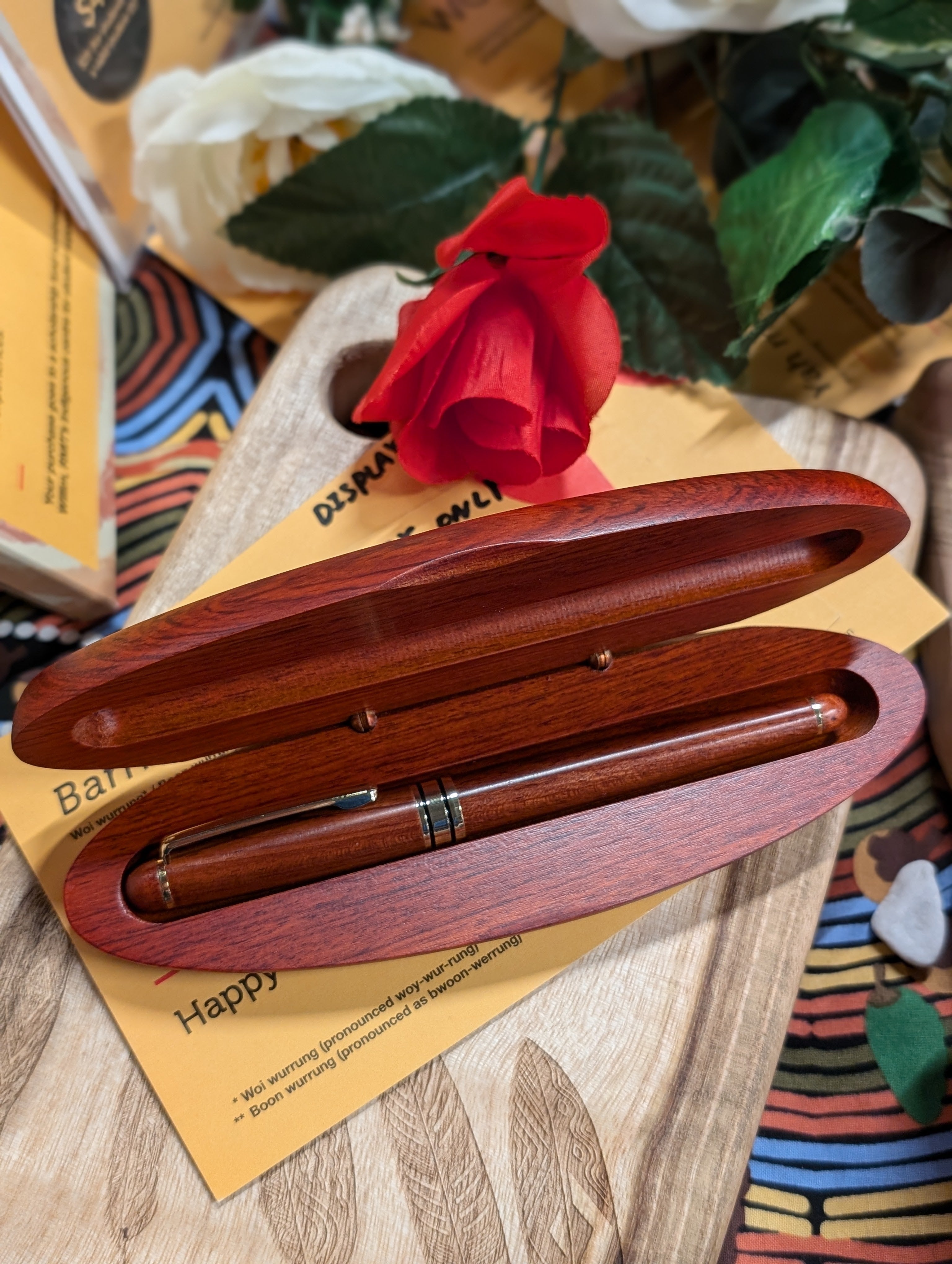 An elegant rosewood pen set from the RMIT Store displayed on a carved wooden surface with Indigenous patterns. The pen set includes a finely crafted pen in a matching rosewood case, surrounded by vibrant flowers and educational cards, reflecting the cultural context.