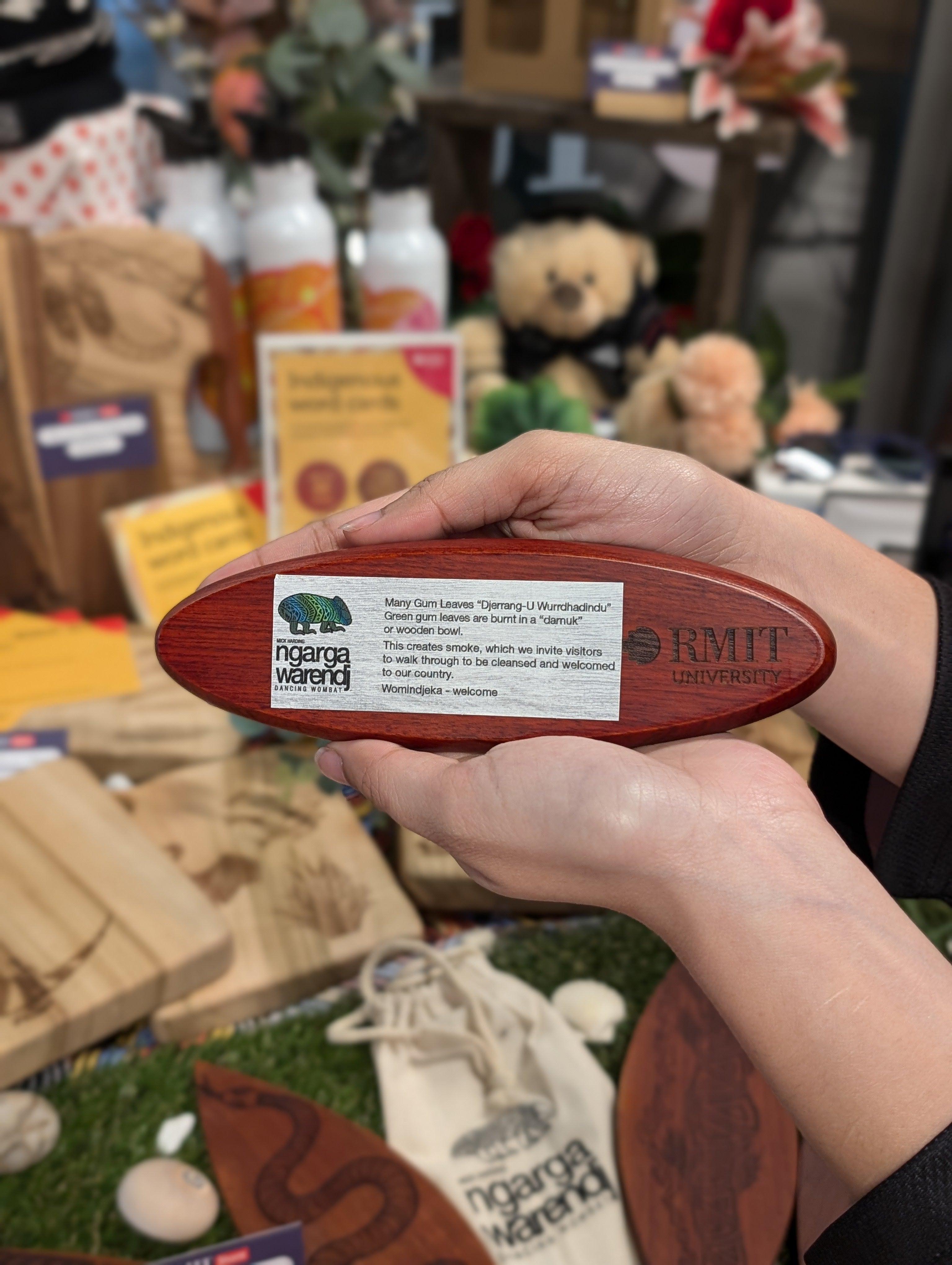 Hands holding a rosewood pen set from the RMIT Store, showcasing engraved branding from Ngarga Warendj and RMIT University. The set features a description card about cultural significance, amidst a backdrop of Indigenous-themed products, highlighting authenticity and artistry.