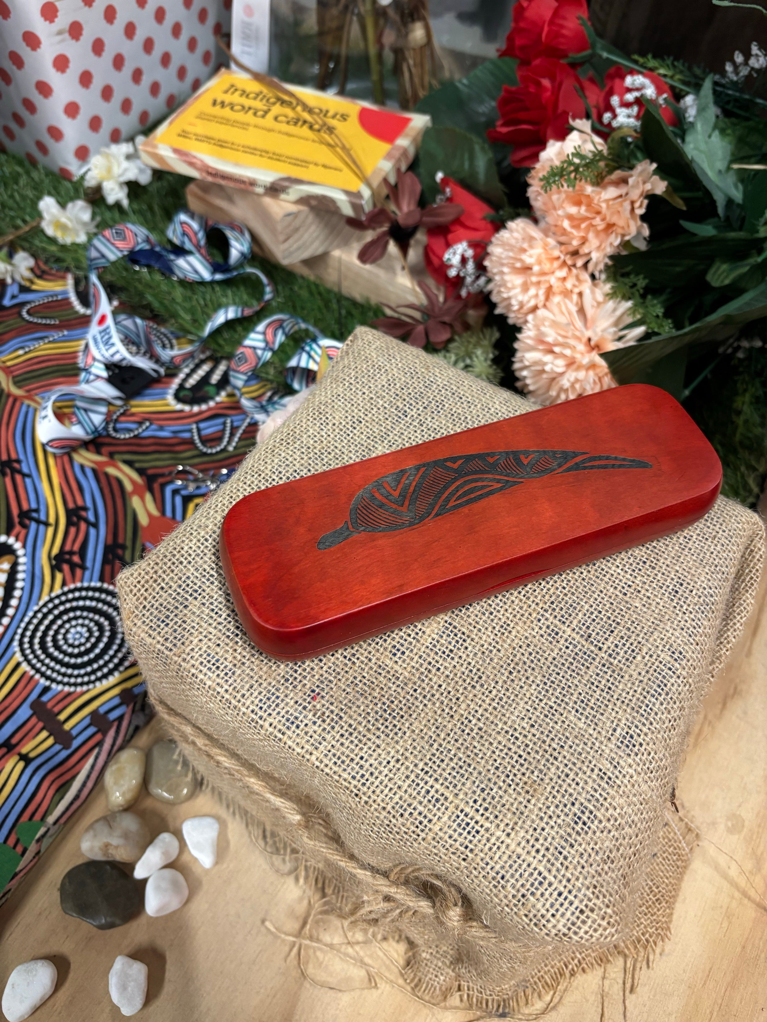 Rosewood Rectangular Pen Set with smooth polished red-brown wooden finish, featuring a black engraved gum leaf motif centred on the lid, displayed closed on a hessian-wrapped box with decorative Aboriginal-inspired patterned fabric, assorted small stones, flowers, and Indigenous word cards in the background, available at the RMIT Store.