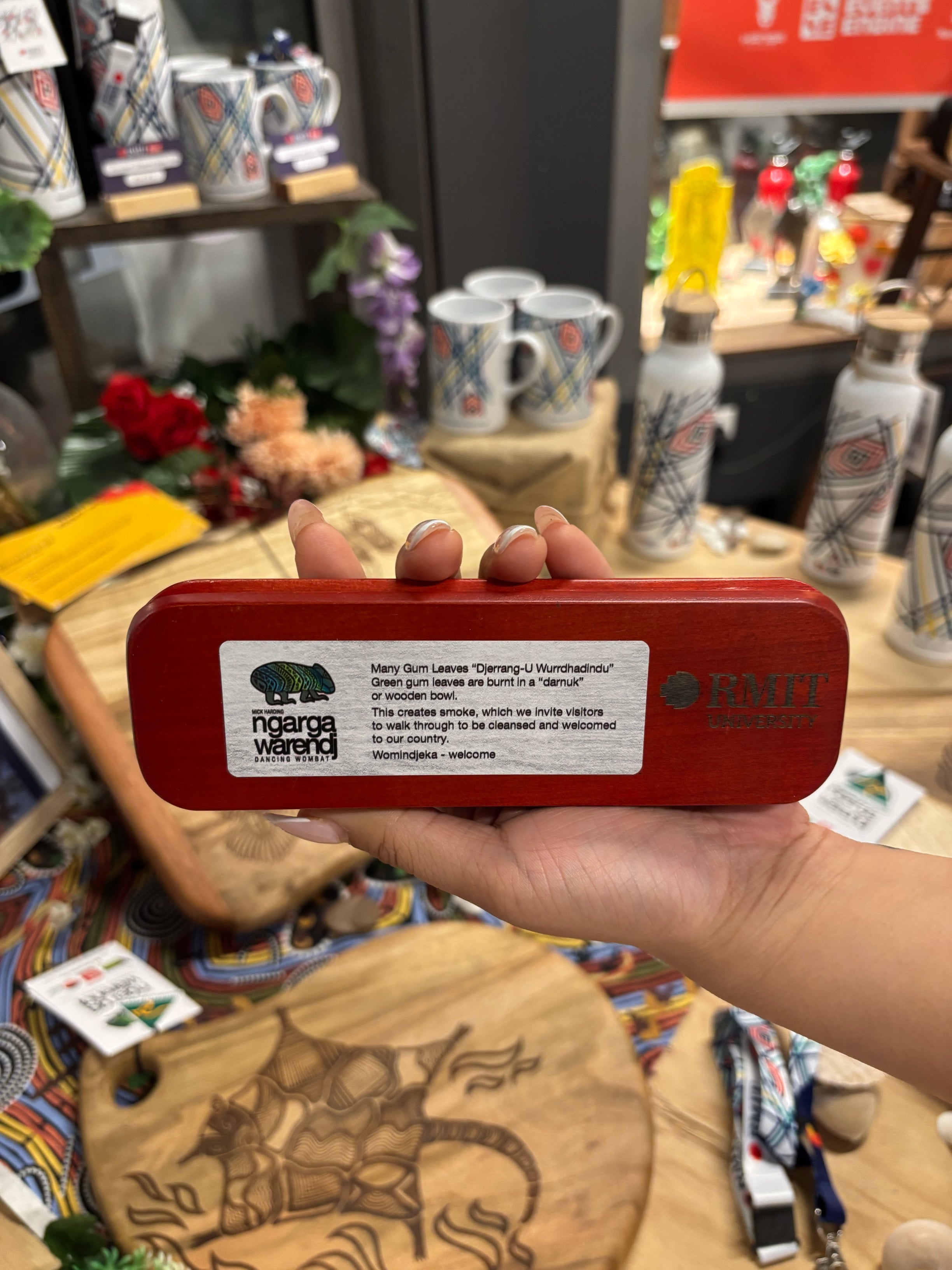 Rosewood Rectangular Pen Set held by a model, showing the underside engraved with Ngarga Warendj Dancing Wombat logo, RMIT University logo, and a printed Welcome to Country message describing the cultural significance of burning green gum leaves, displayed in-store with Aboriginal-inspired patterned fabric, engraved wooden serving boards, ceramic mugs, drink bottles, and floral arrangements in the background, available at the RMIT Store.