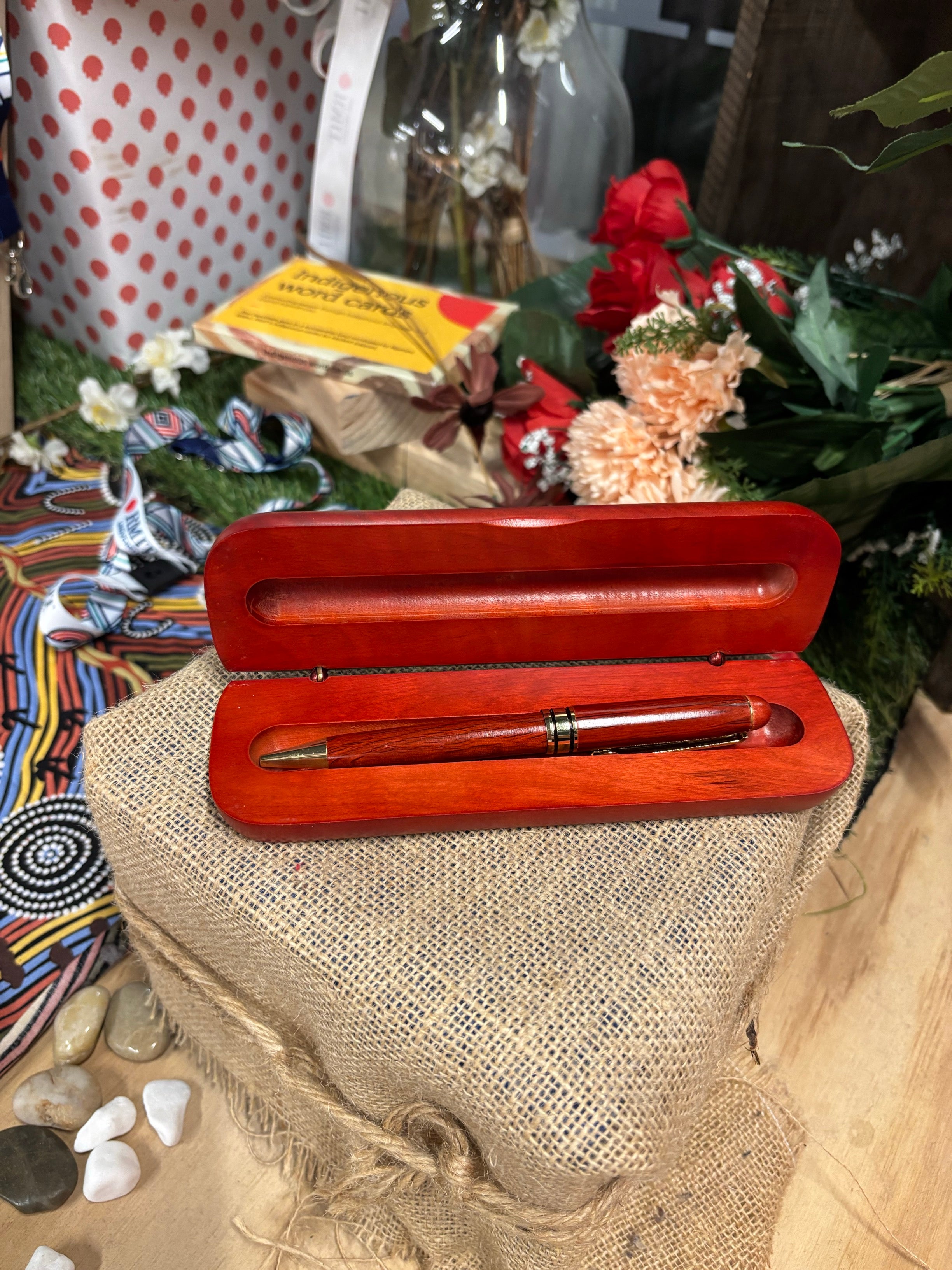 Rosewood Rectangular Pen Set with smooth polished red-brown wooden case, opened to reveal a matching rosewood pen with gold trim nestled inside, displayed on a hessian-wrapped box with decorative Aboriginal-inspired patterned fabric, assorted small stones, flowers, and Indigenous word cards in the background, available at the RMIT Store.