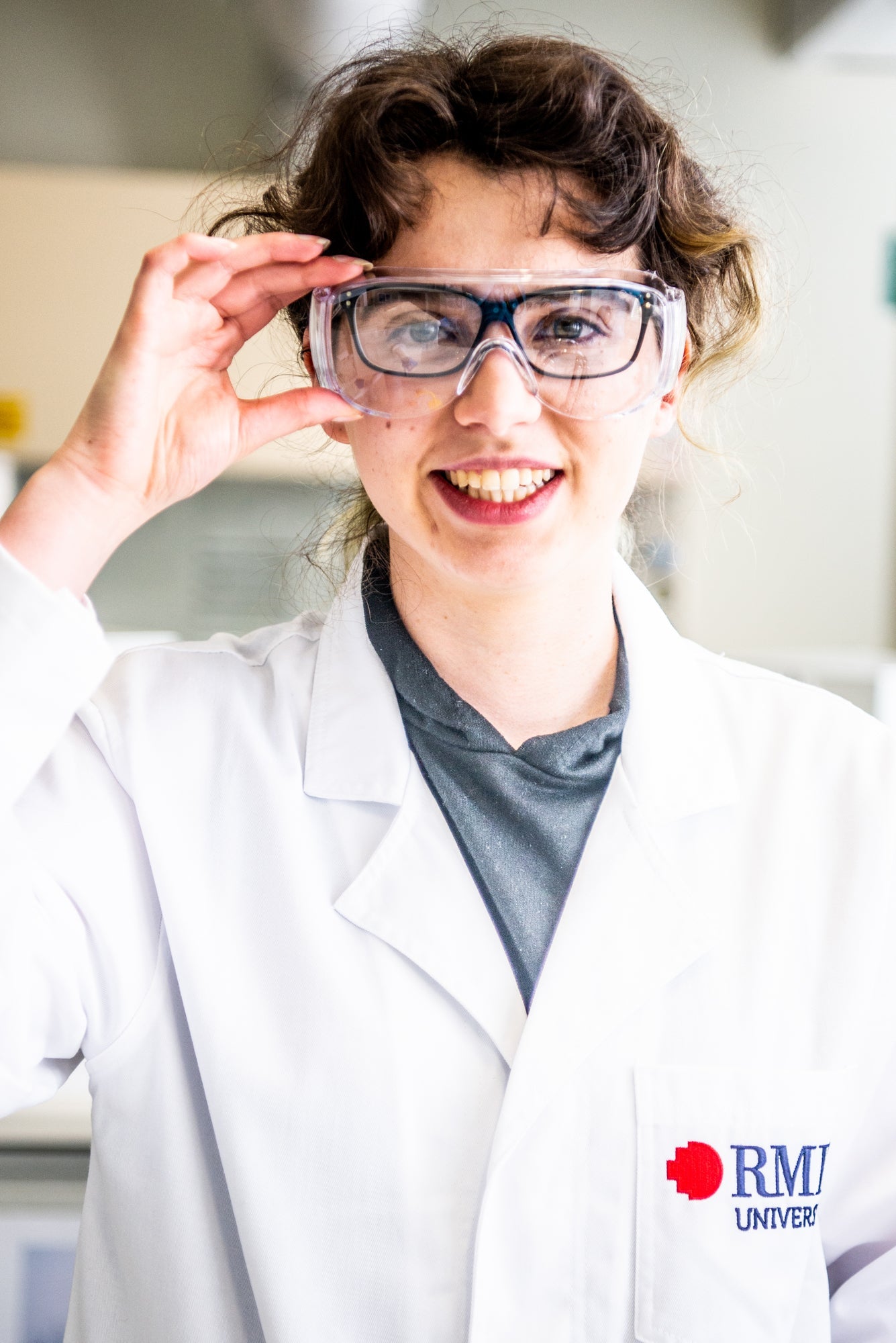 Model adjusting safety glasses while wearing a white RMIT University lab coat, demonstrating laboratory safety equipment.