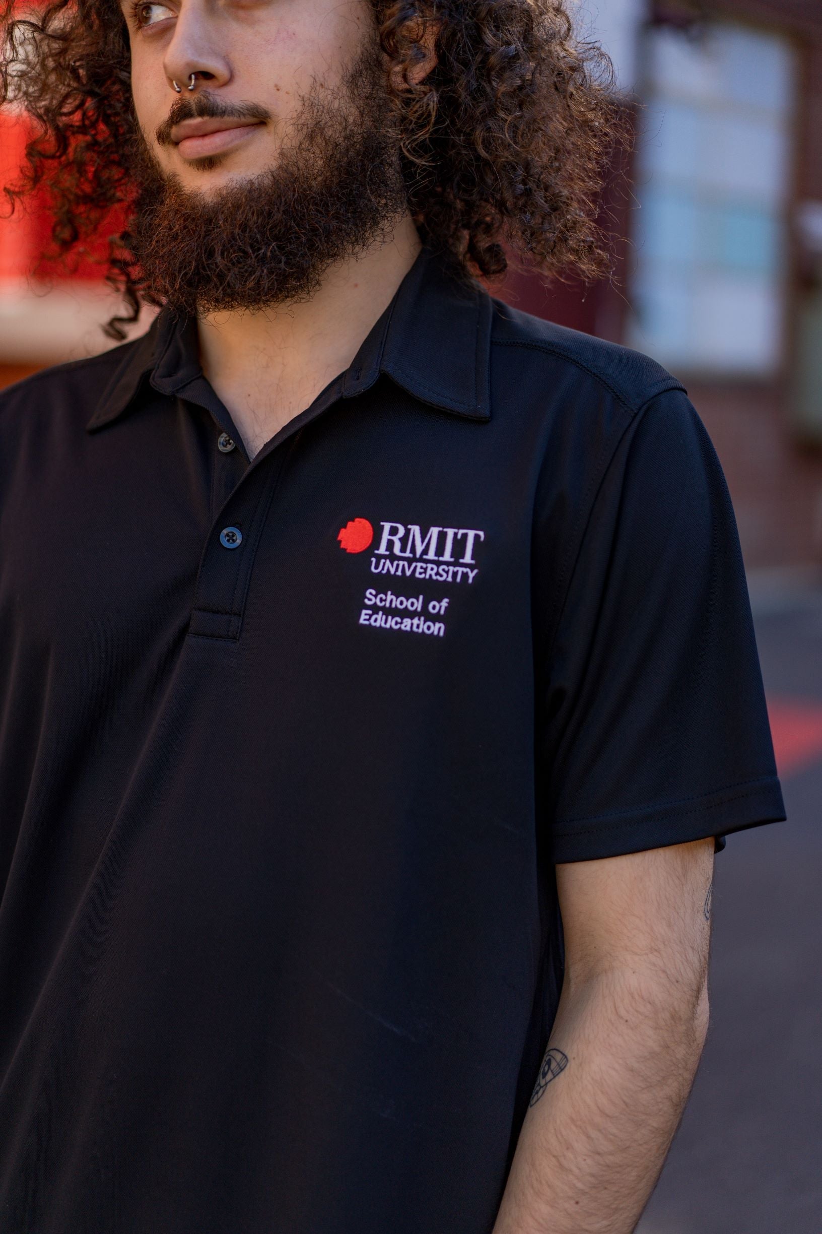 Male model wearing the RMIT School of Education Polo in black, photographed front-on outdoors on the RMIT campus with buildings and paved pathways visible in the background. The short-sleeved polo features “School of Education” embroidered in white on the right chest and the RMIT University logo embroidered in red and white on the left chest. 