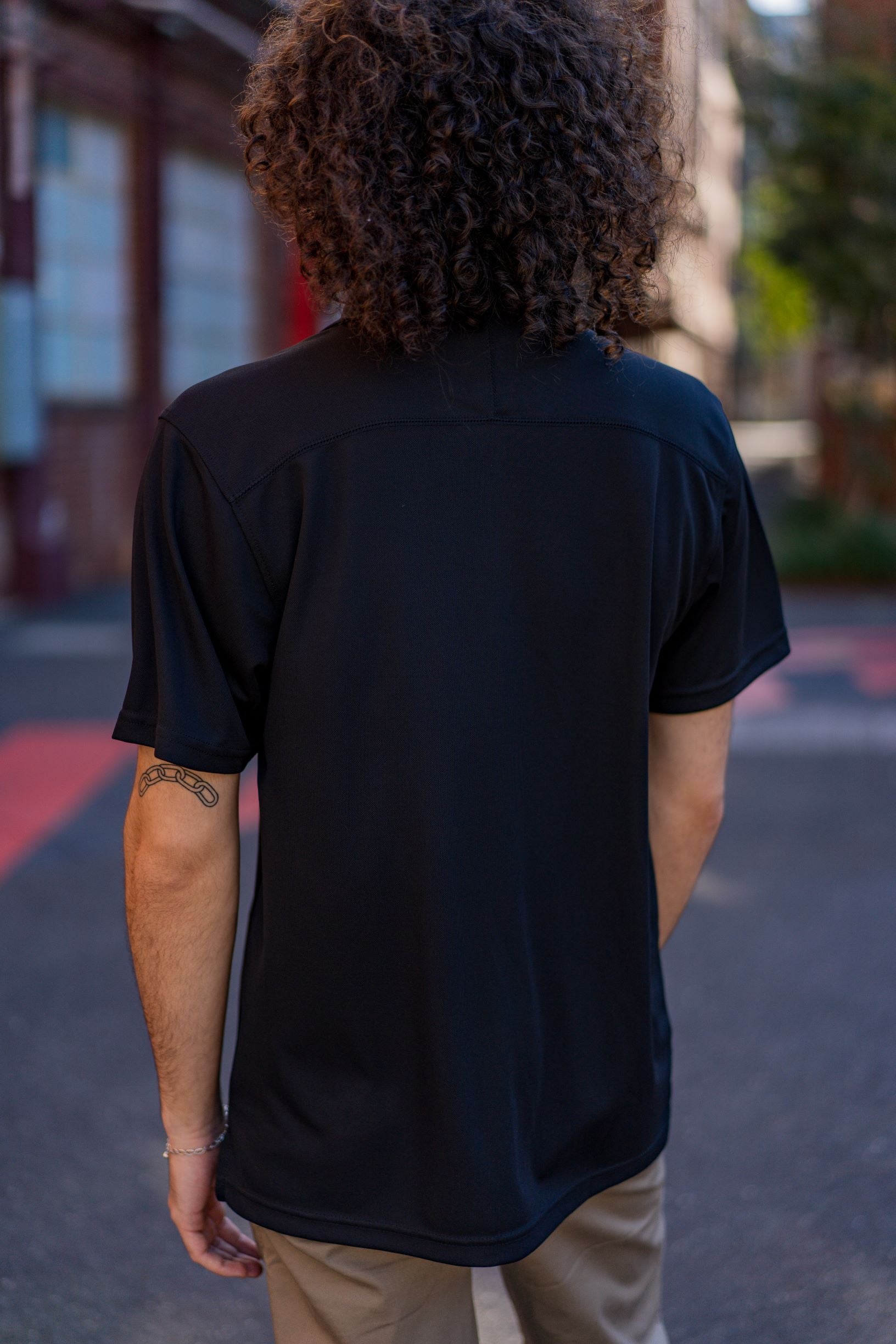 Rear view of a male model wearing the RMIT School of Education Polo in black, standing outdoors on the RMIT campus with buildings and paved pathways visible in the background. The model stands with arms relaxed by his sides, shown in natural daylight for the RMIT Store.