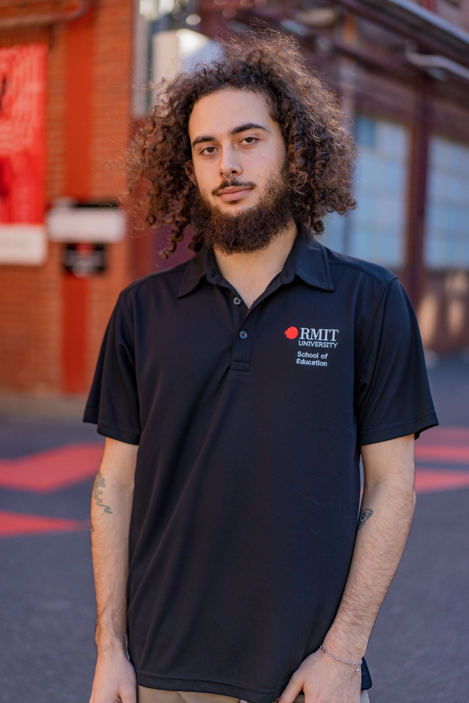 Male model wearing the RMIT School of Education Polo in black, photographed front-on outdoors on the RMIT campus with buildings and paved pathways visible in the background. The short-sleeved polo features “School of Education” embroidered in white on the right chest and the RMIT University logo embroidered in red and white on the left chest. The model stands with arms relaxed by his sides, shown in natural daylight for the RMIT Store.