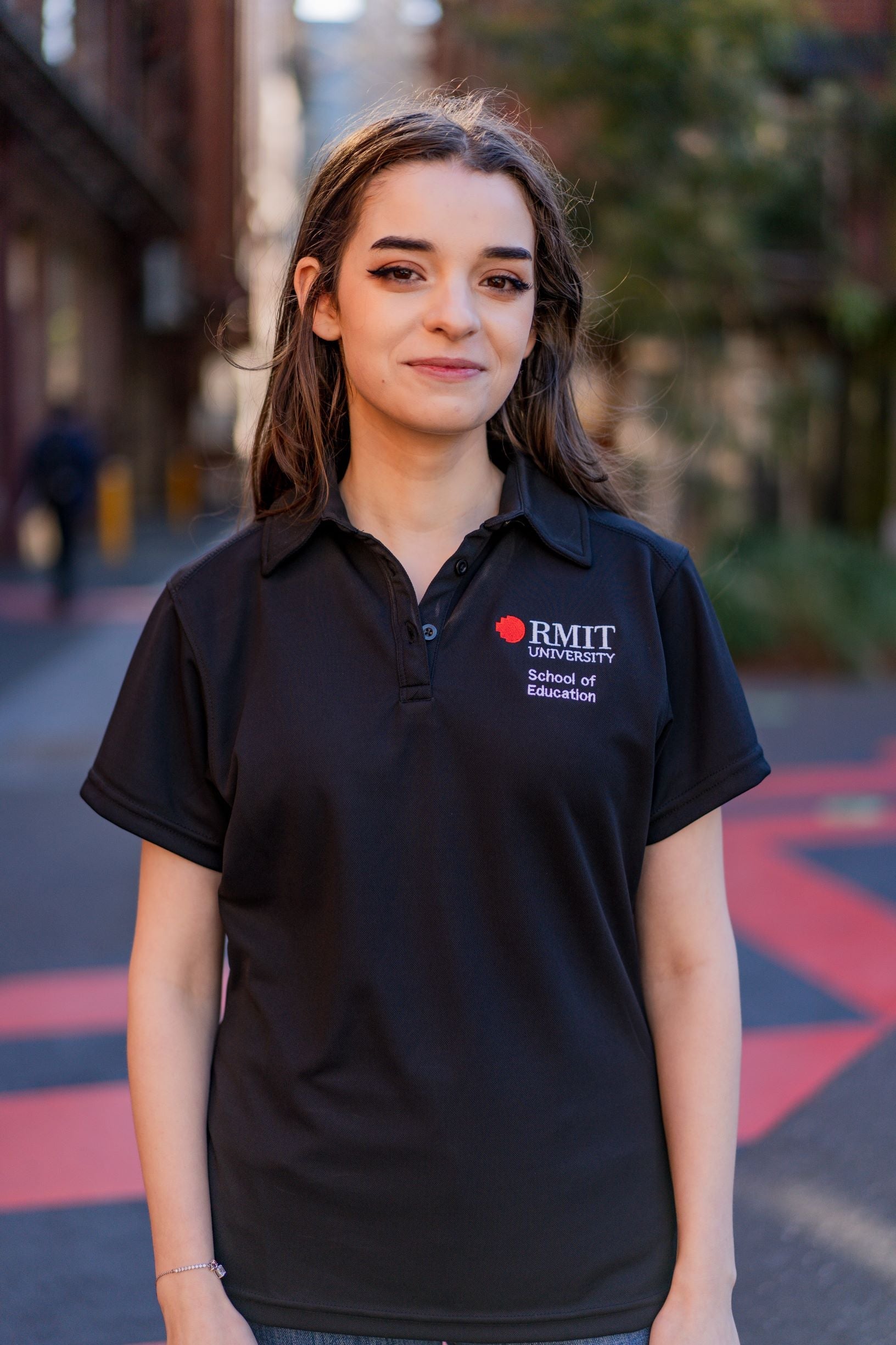 Model wearing the RMIT School of Education Polo in black, photographed front-on outdoors on the RMIT campus with buildings and paved pathways visible in the background. The short-sleeved polo features “School of Education” embroidered in white on the right chest and the RMIT University logo embroidered in red and white on the left chest. The model stands with arms relaxed by their sides, shown in natural daylight for the RMIT Store.