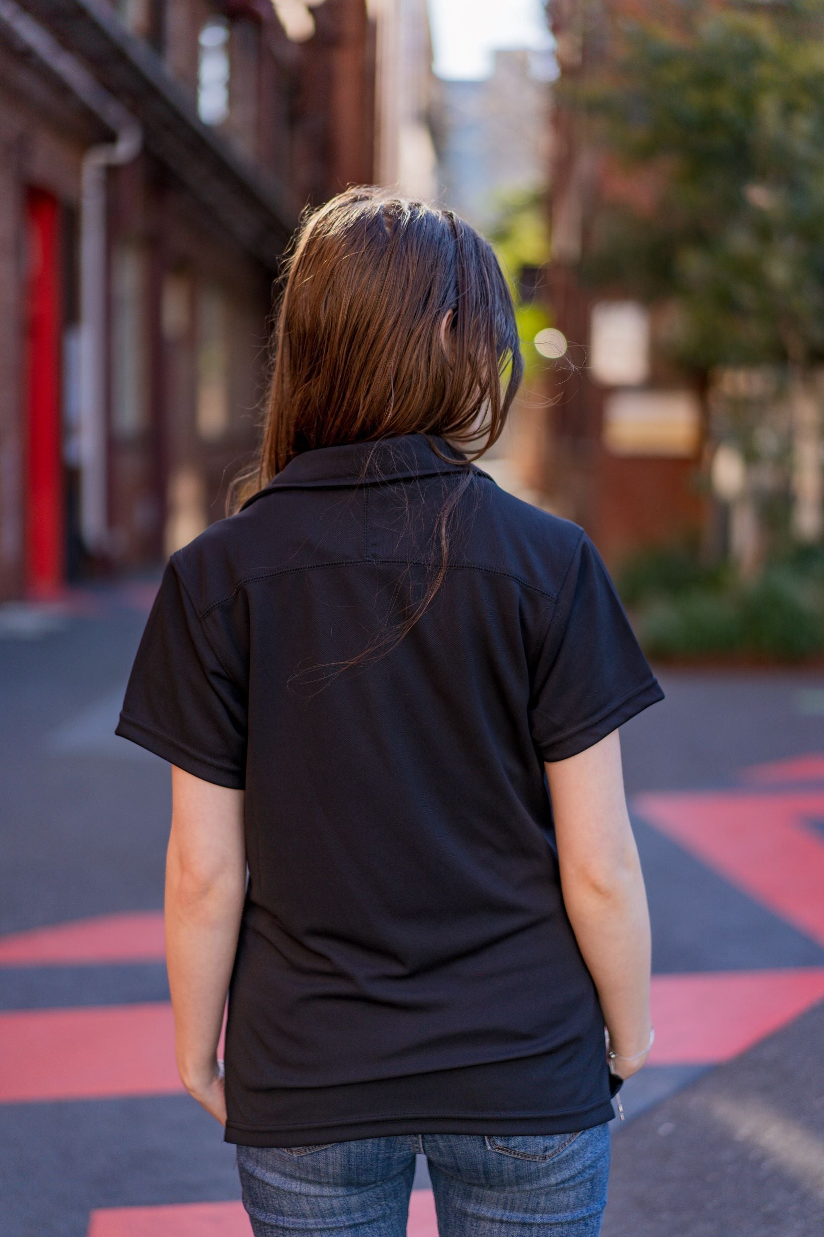 Rear view of the RMIT School of Education Polo in black worn by a model standing outdoors on the RMIT campus with buildings and paved pathways visible in the background. The model is slightly turned to the right, showcasing the plain black back of the short-sleeved polo. Both arms are relaxed by her sides. Image captured in natural daylight for the RMIT Store.