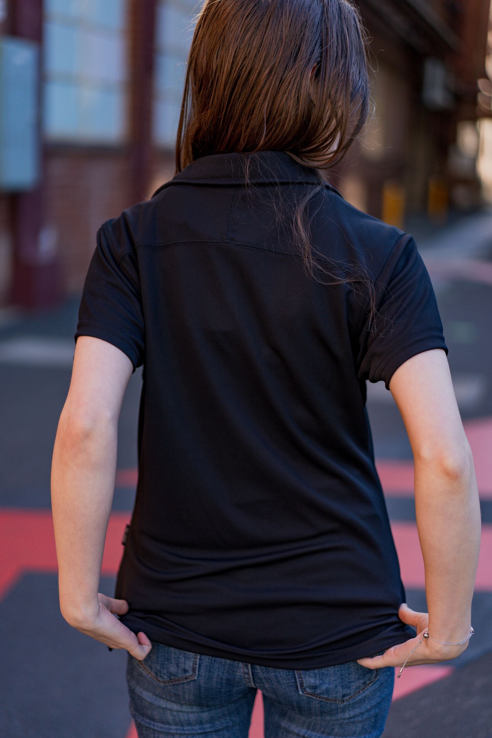 Rear view of the RMIT School of Education Polo in black worn by a model standing outdoors on the RMIT campus with buildings and paved pathways in the background. The model holds the bottom edge of the short-sleeved polo with both hands, slightly lifting the fabric. Natural daylight highlights the product for the RMIT Store.